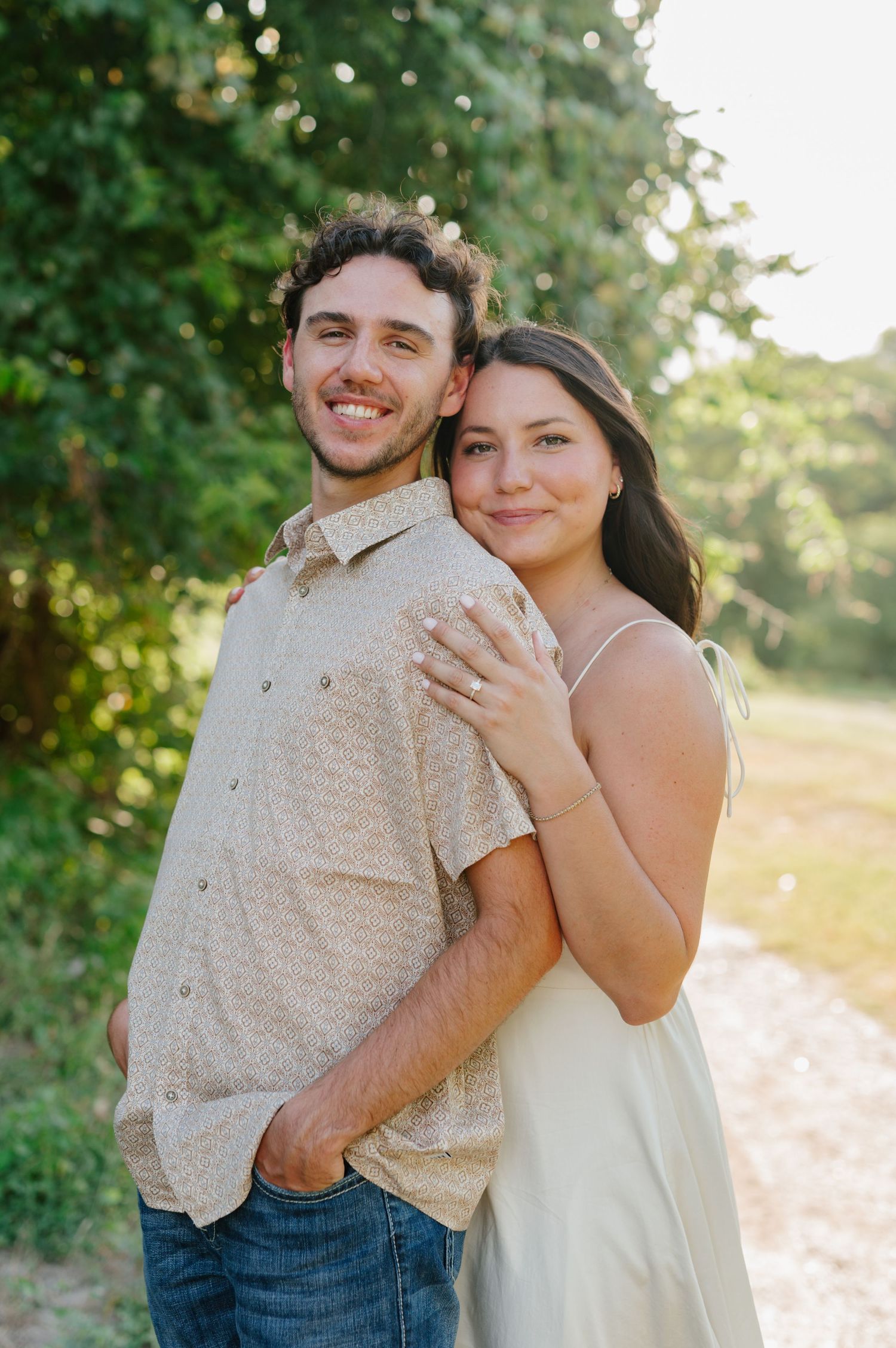 A couple embraces in a romantic outdoor photoshoot with golden sunlight filtering through trees in the background.