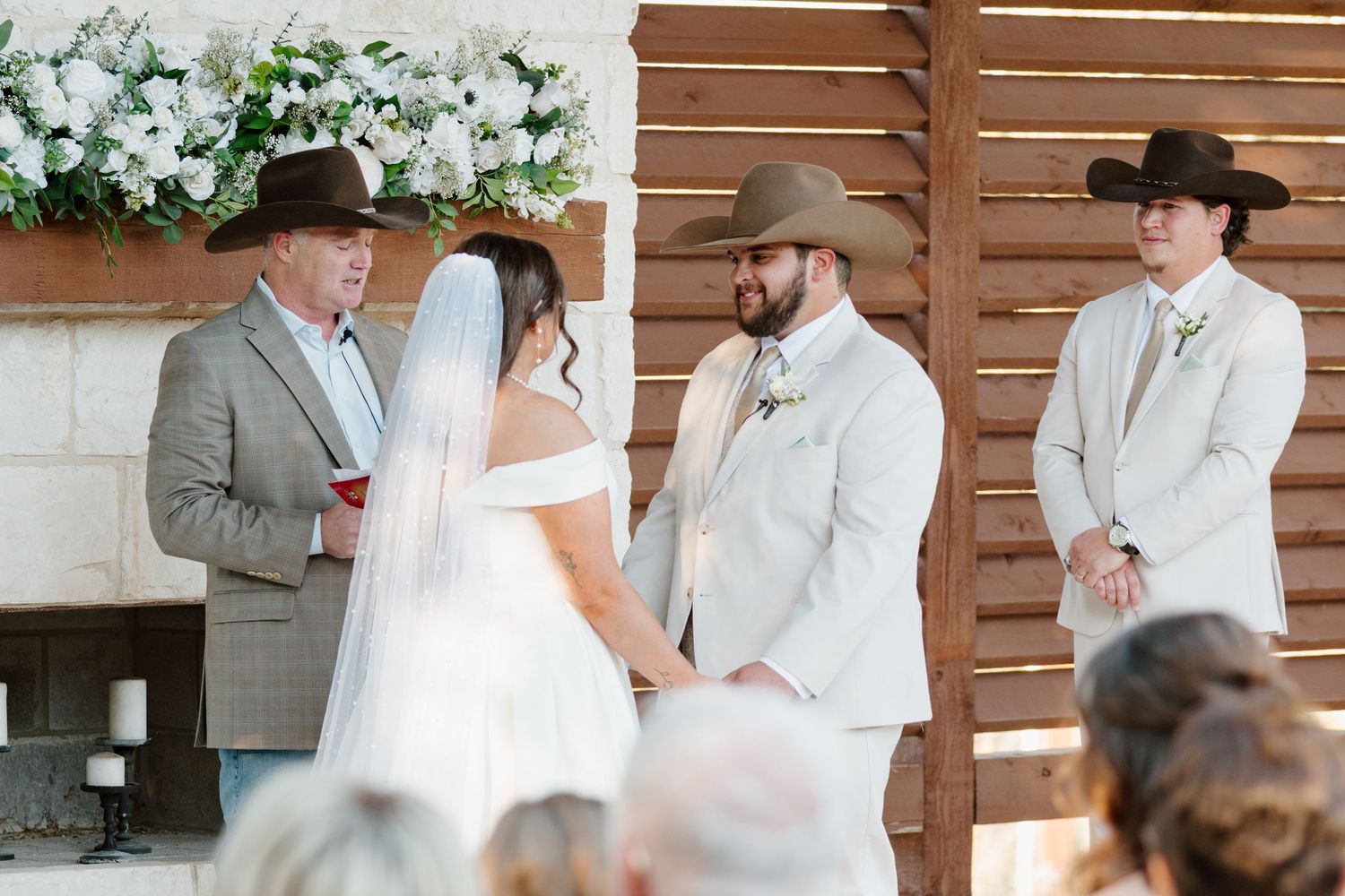 A wedding ceremony takes place outdoors with people in cowboy hats and formal attire standing at a wooden backdrop.