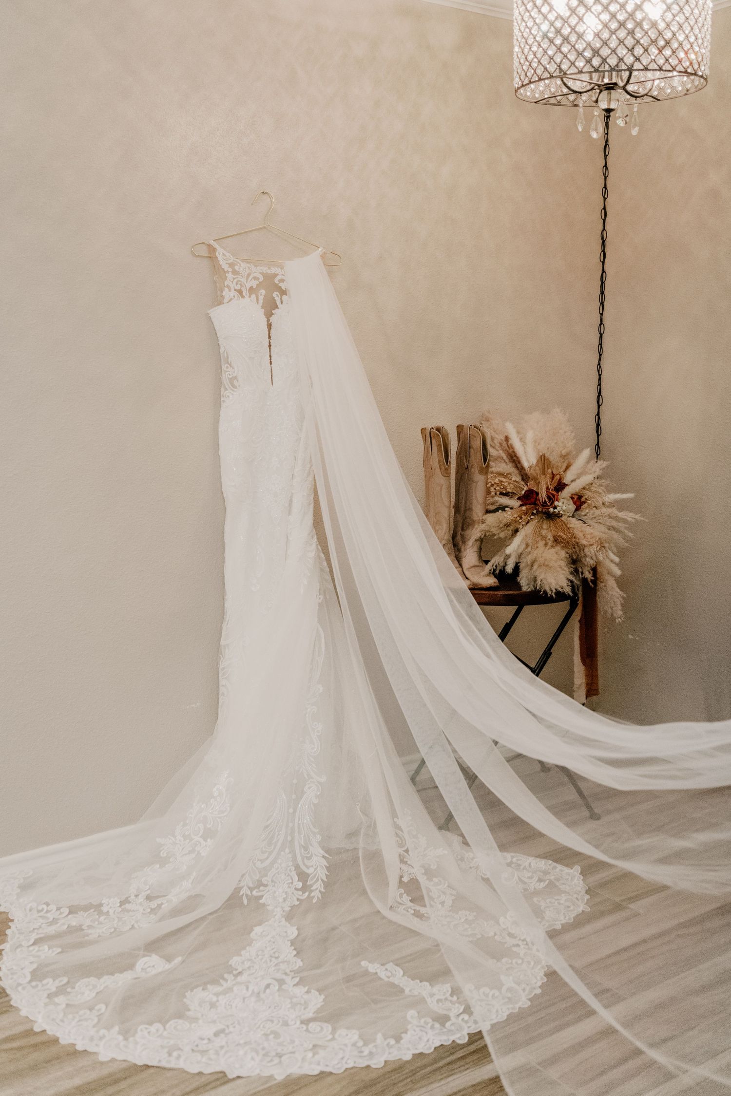 Beautiful white lace wedding dress with long trailing veil displayed next to a floor lamp and dried flower arrangement.