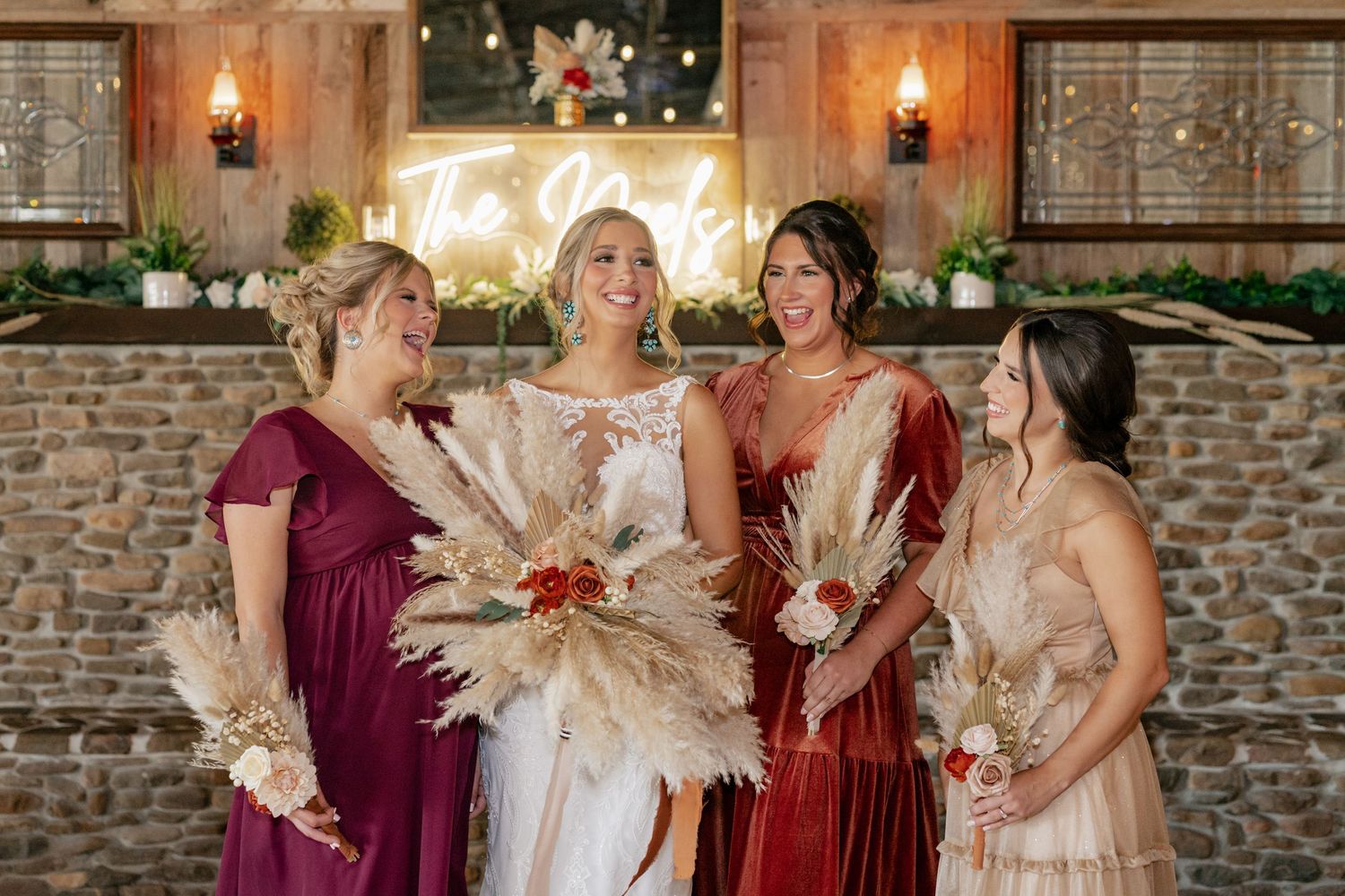 Wedding party wearing burgundy and champagne dresses holding dried pampas grass bouquets against stone wall.