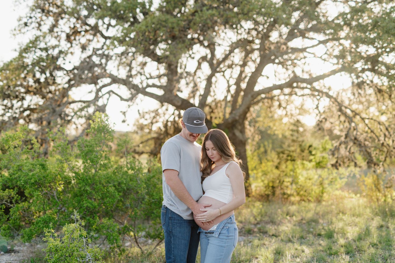Couple embraces in golden sunlight among trees and greenery during sunset maternity photo session.