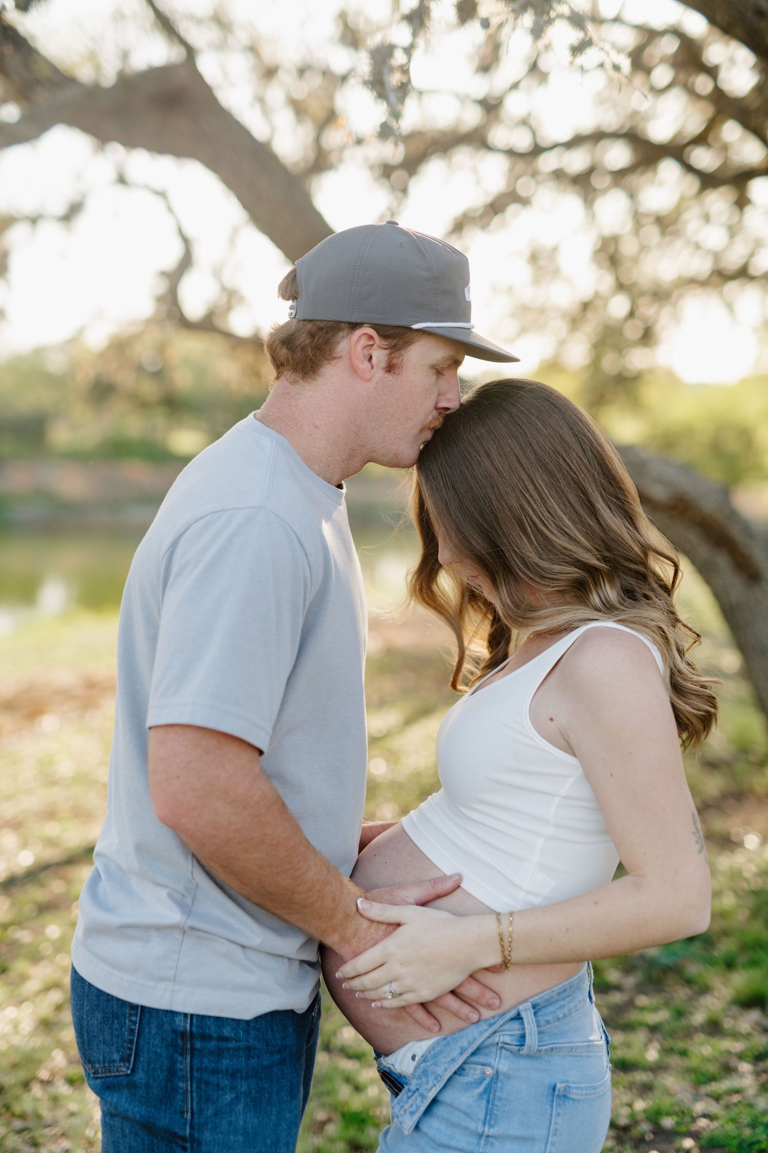 Expecting couple shares tender moment under sunlit trees during outdoor maternity photoshoot.