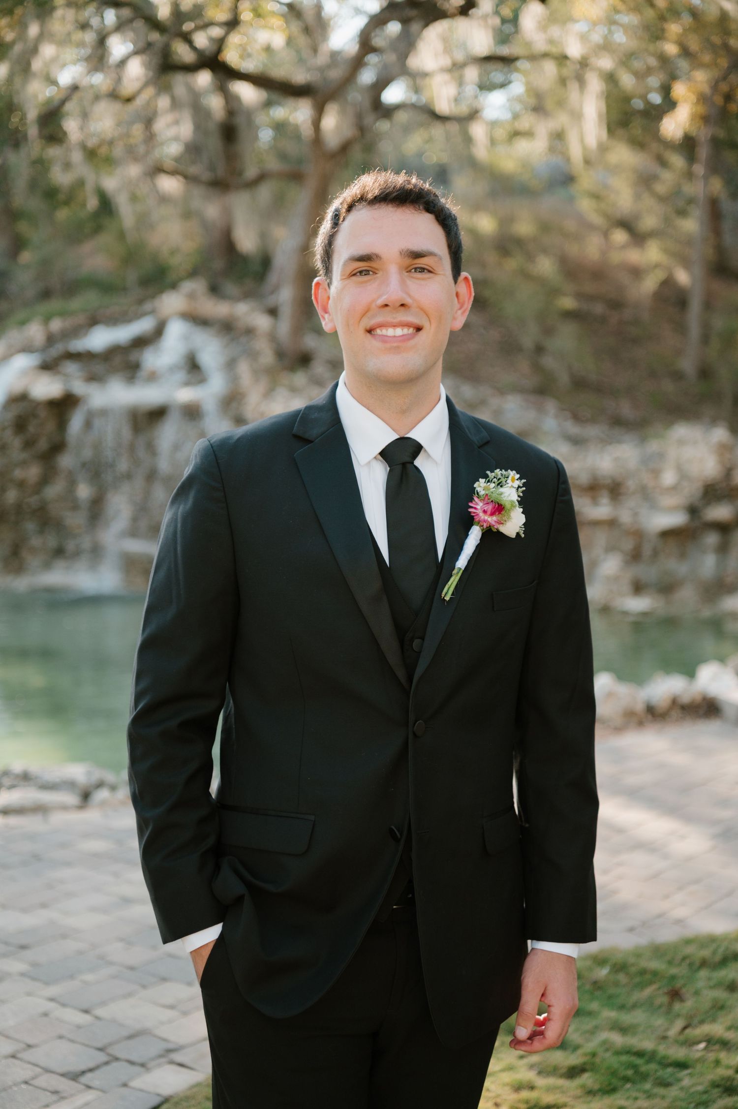 Groom in classic black suit and tie poses outdoors against natural stone backdrop.