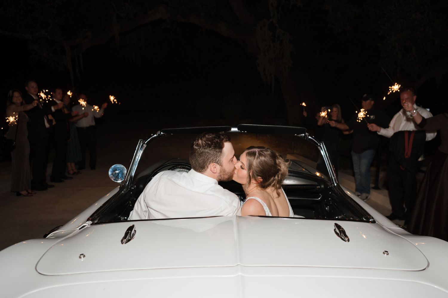 A couple shares a romantic moment in a vintage white car while guests hold sparklers around them photographed by Alyssa Denson in Texas.