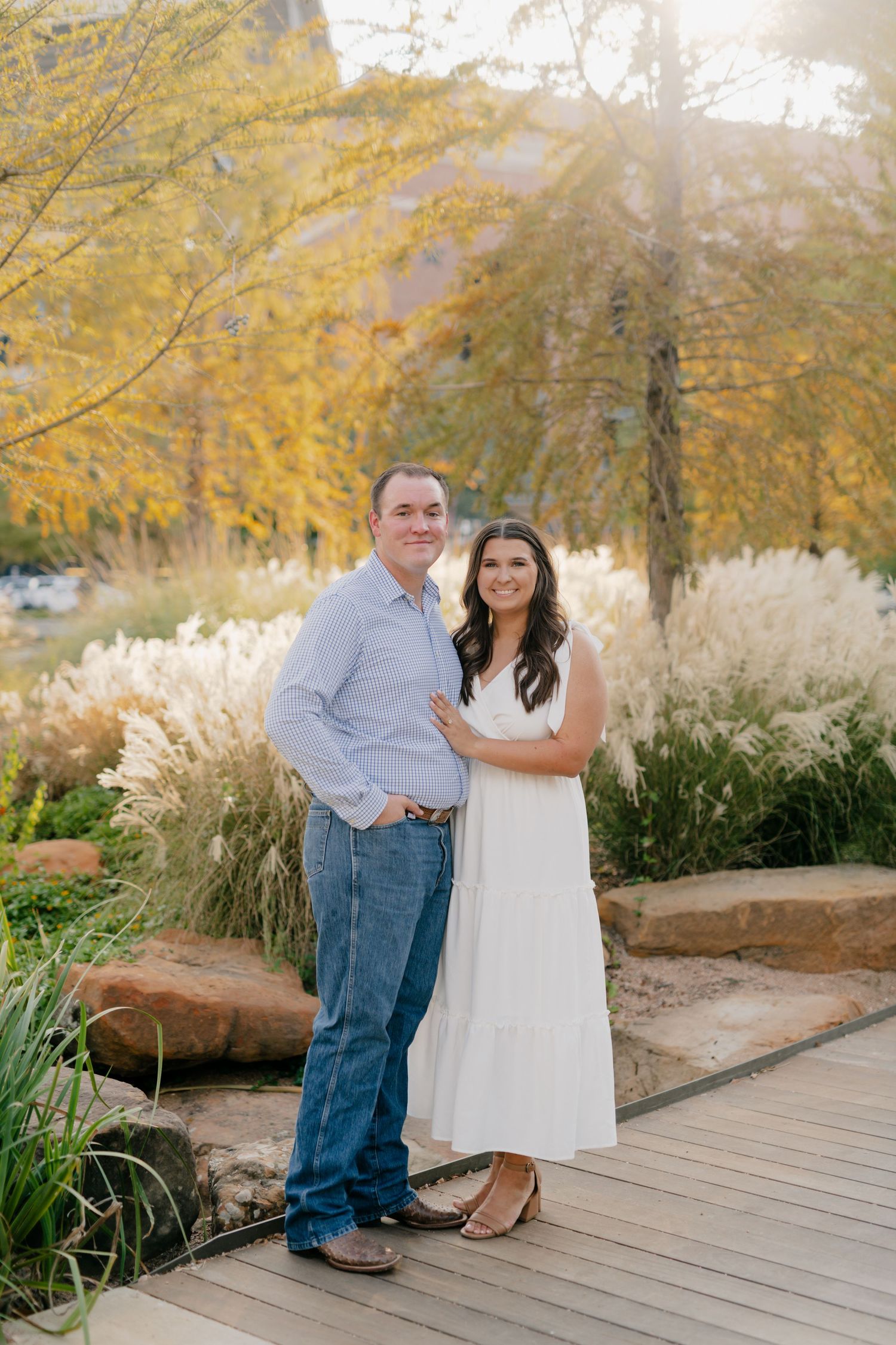Couple stands on wooden deck surrounded by golden autumn trees and ornamental grasses.
