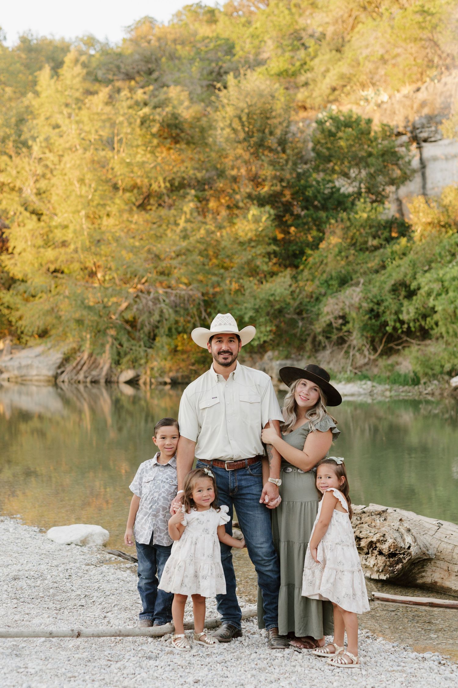 A family enjoys a fall photoshoot by a tranquil river with autumn foliage in the background.
