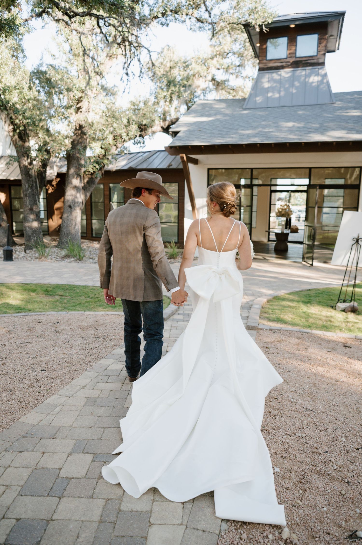 A bride in a flowing white dress walks with her partner along a stone path surrounded by trees.