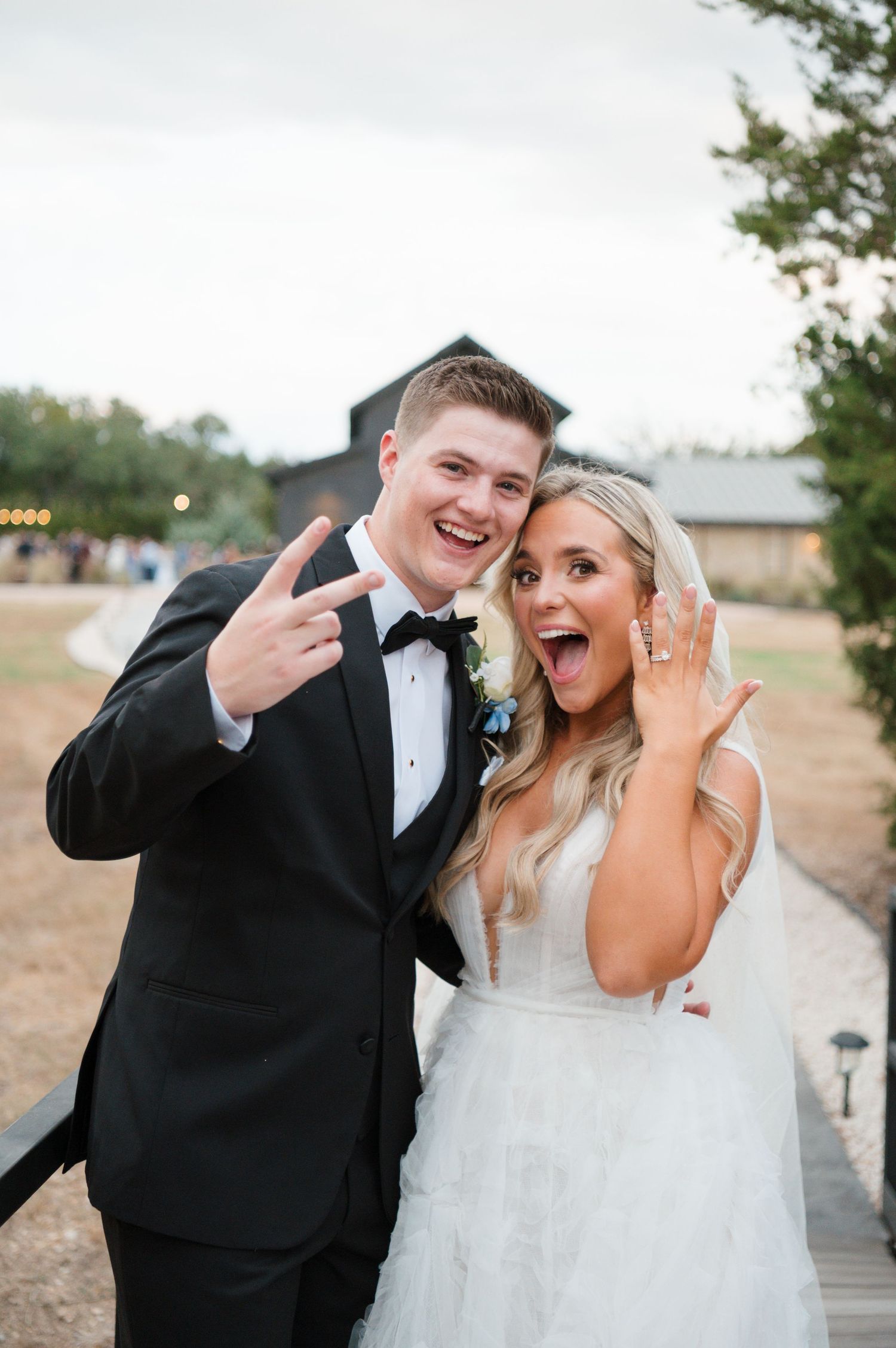 A bride and groom smiling and making a hand gesture while standing on a wooden walkway.