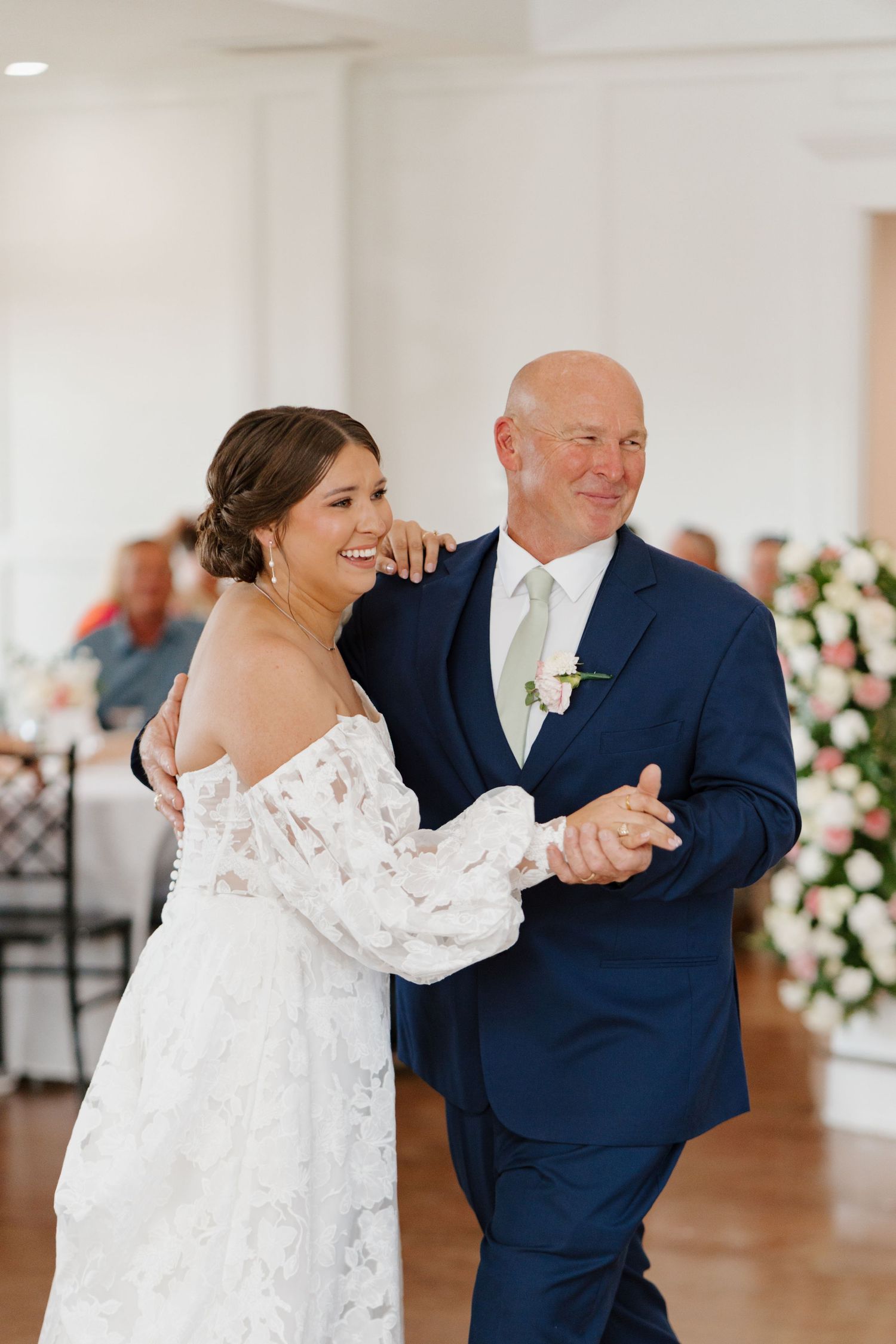 A couple share a playful and intimate dance moment in a bright wedding reception hall decorated with flowers.