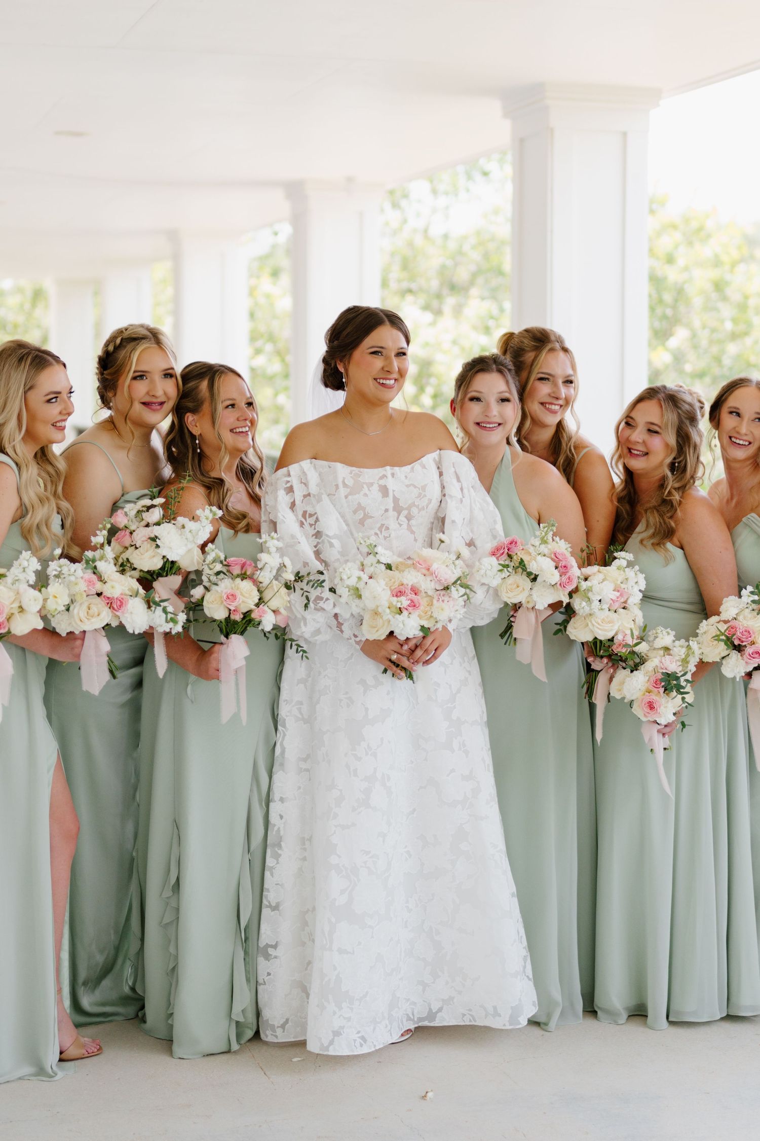 Wedding party in sage green bridesmaid dresses lined up on white porch with pink and white floral bouquets.