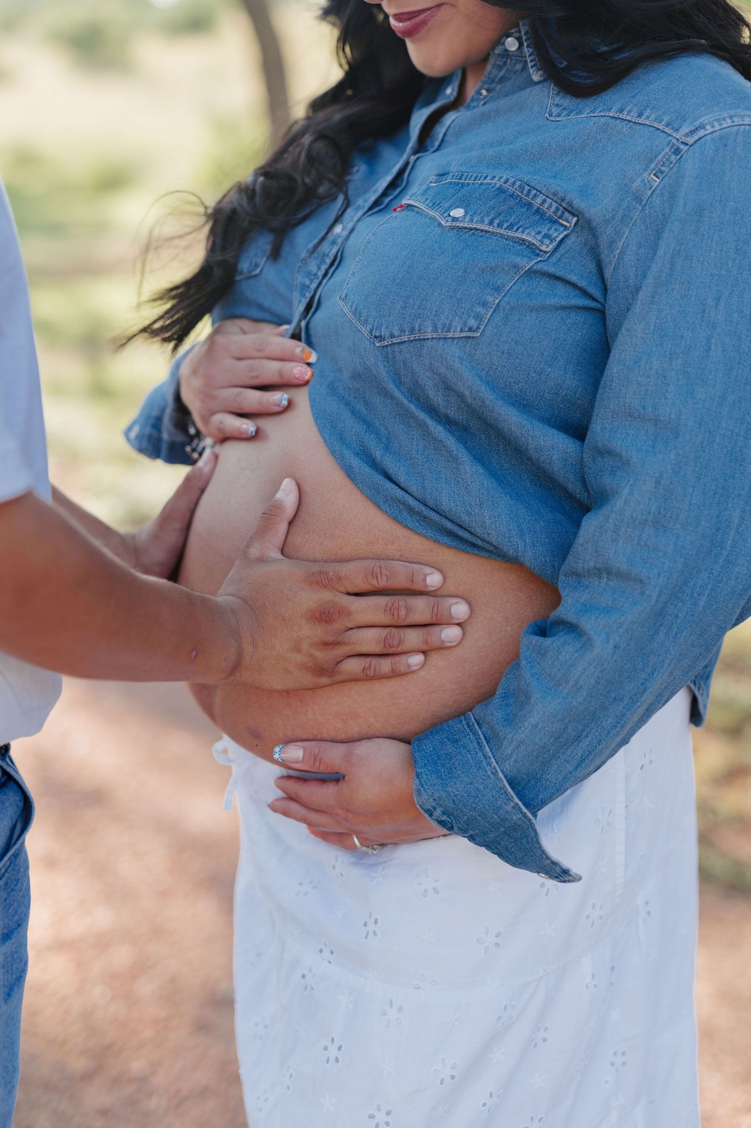 Close-up shot of a pregnant belly being cradled by hands while wearing a denim shirt and white skirt.