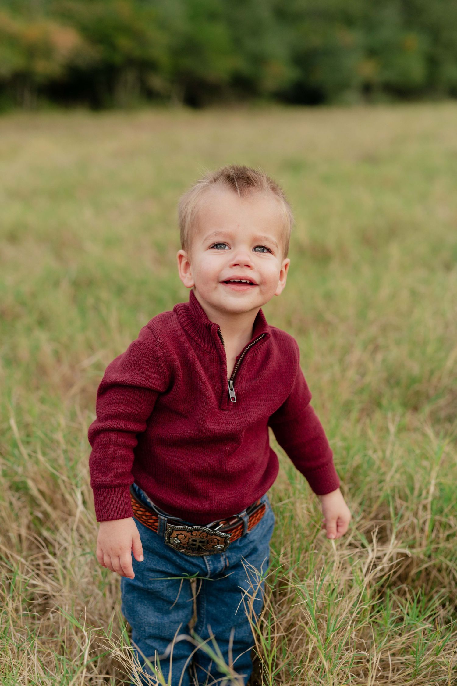 Toddler in burgundy sweater and jeans smiling while standing in tall grass.