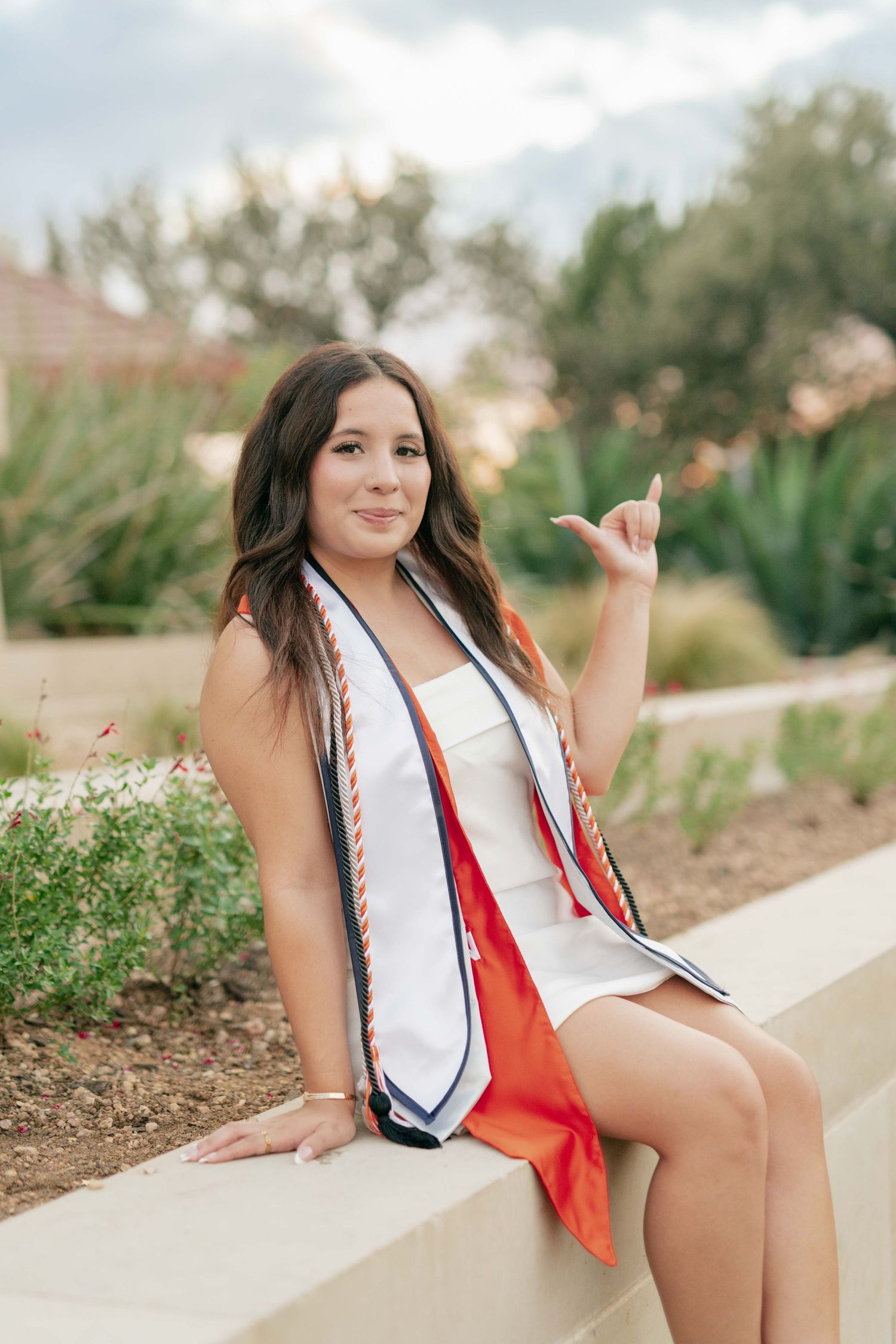 Graduate in white dress and graduation stole makes hand gesture while sitting on concrete wall.