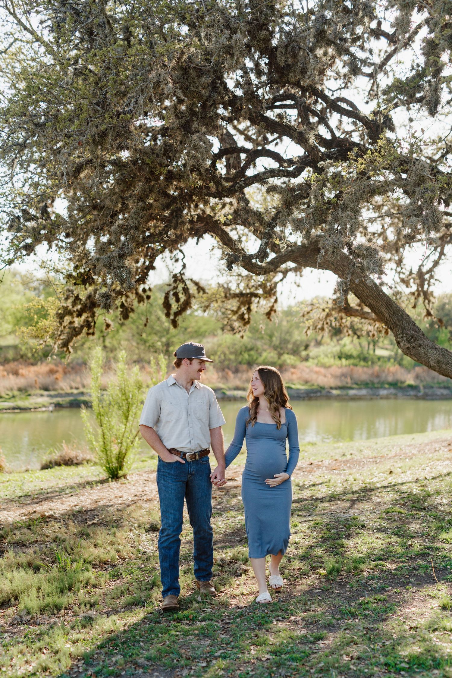 A couple enjoys a romantic walk along a peaceful lake under a large tree in a natural setting.
