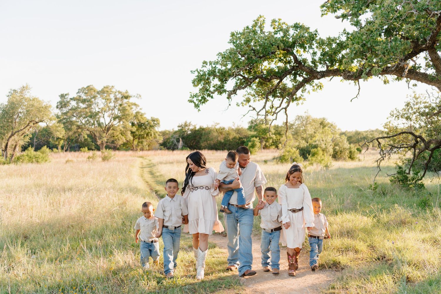A happy family walks together on a tree-lined dirt path through a sunny meadow on a warm afternoon.