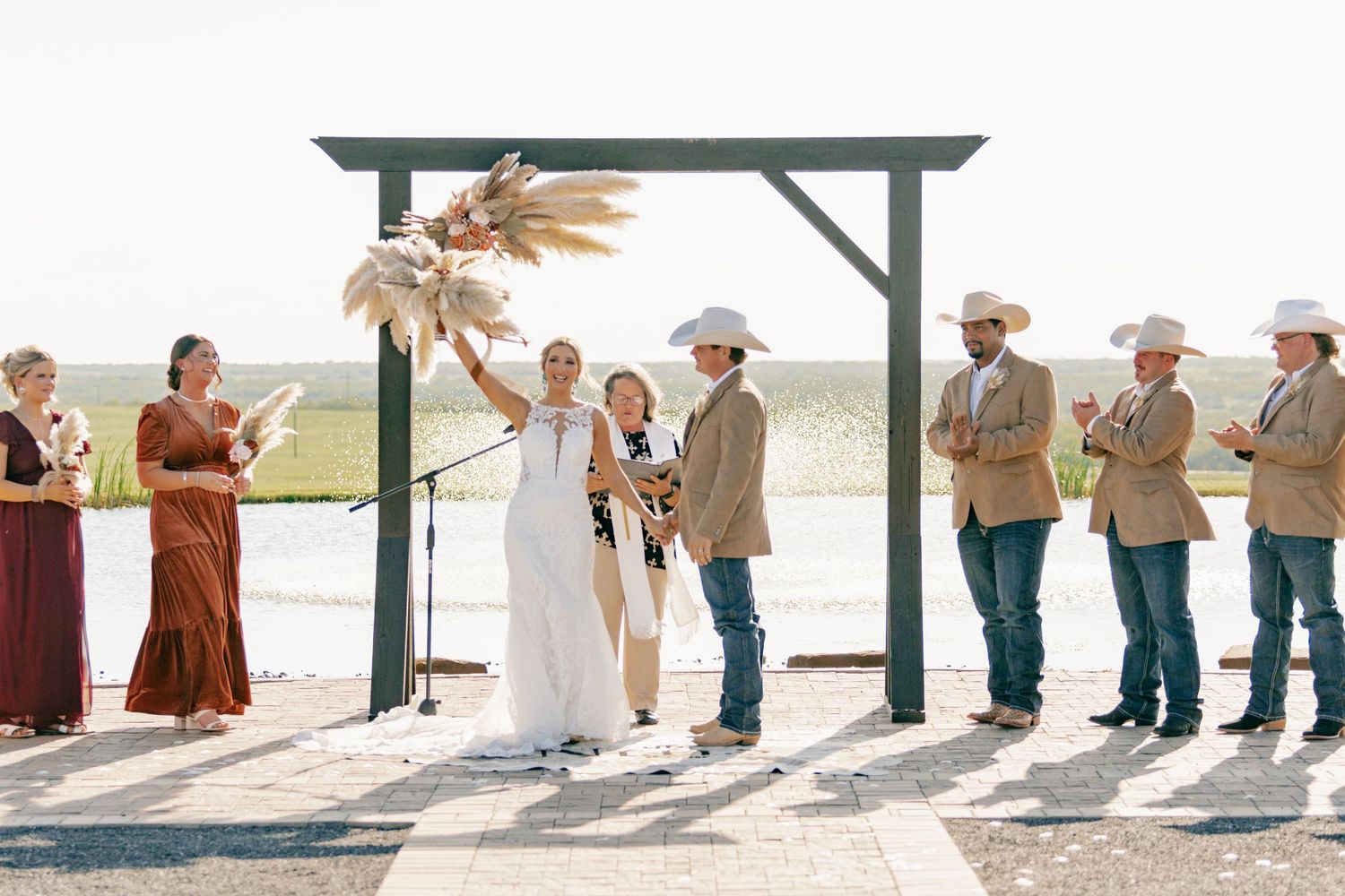 Wedding ceremony under wooden arch with pampas grass and bridal party in burgundy dresses.