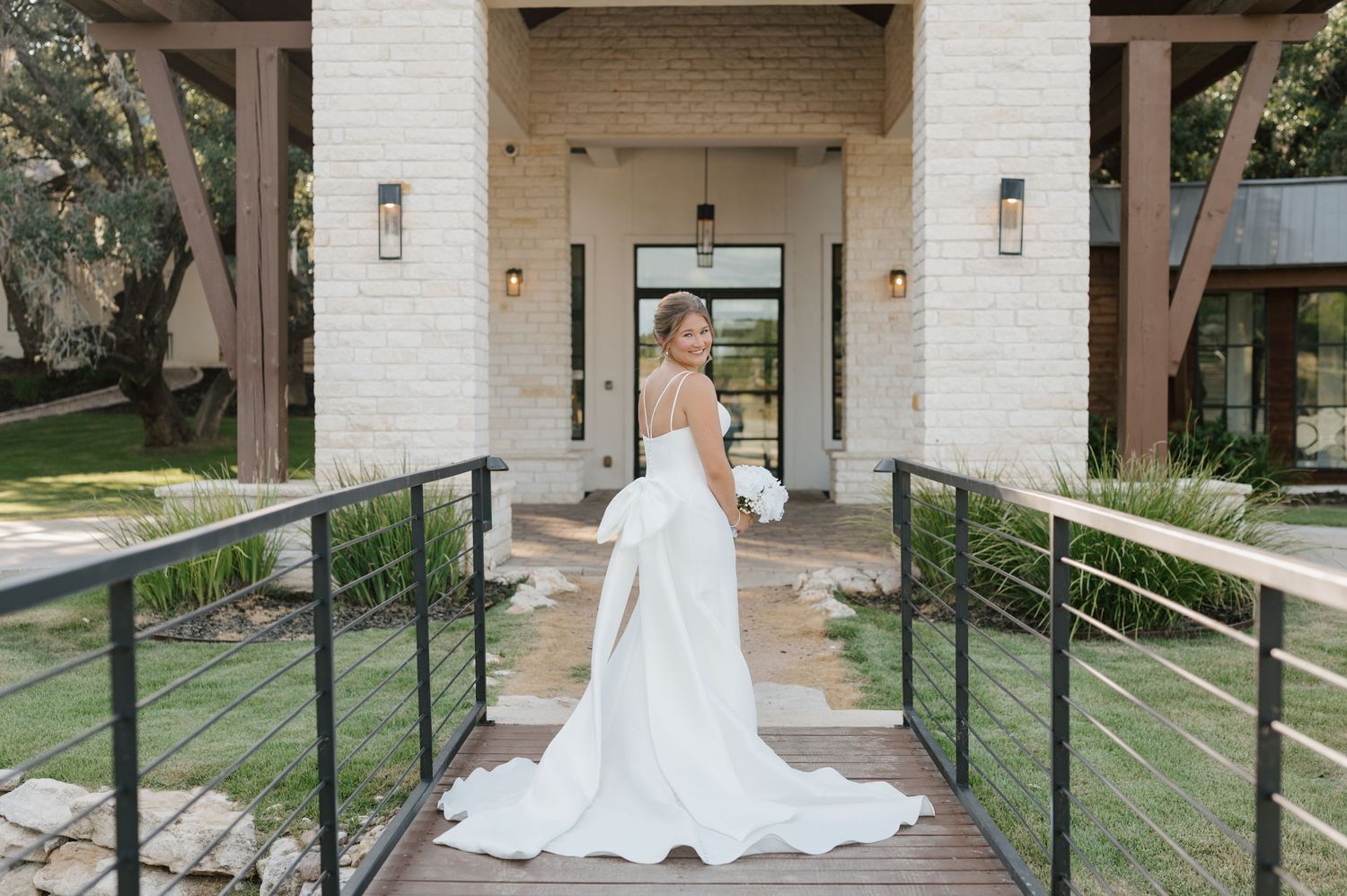 A bride in a flowing white wedding gown poses in an elegant outdoor courtyard with white stone columns and green lawn.