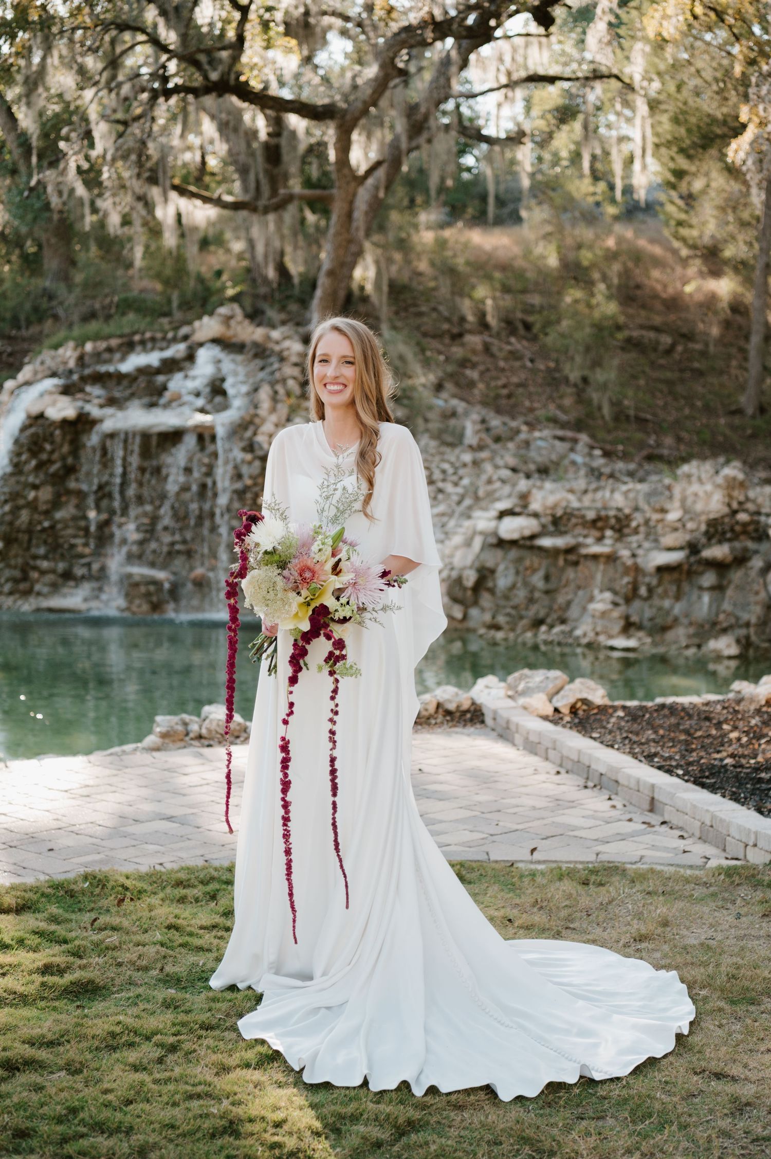 A bride in a long-sleeved white dress holds a stunning bouquet by a scenic lake and rocky cliffs.