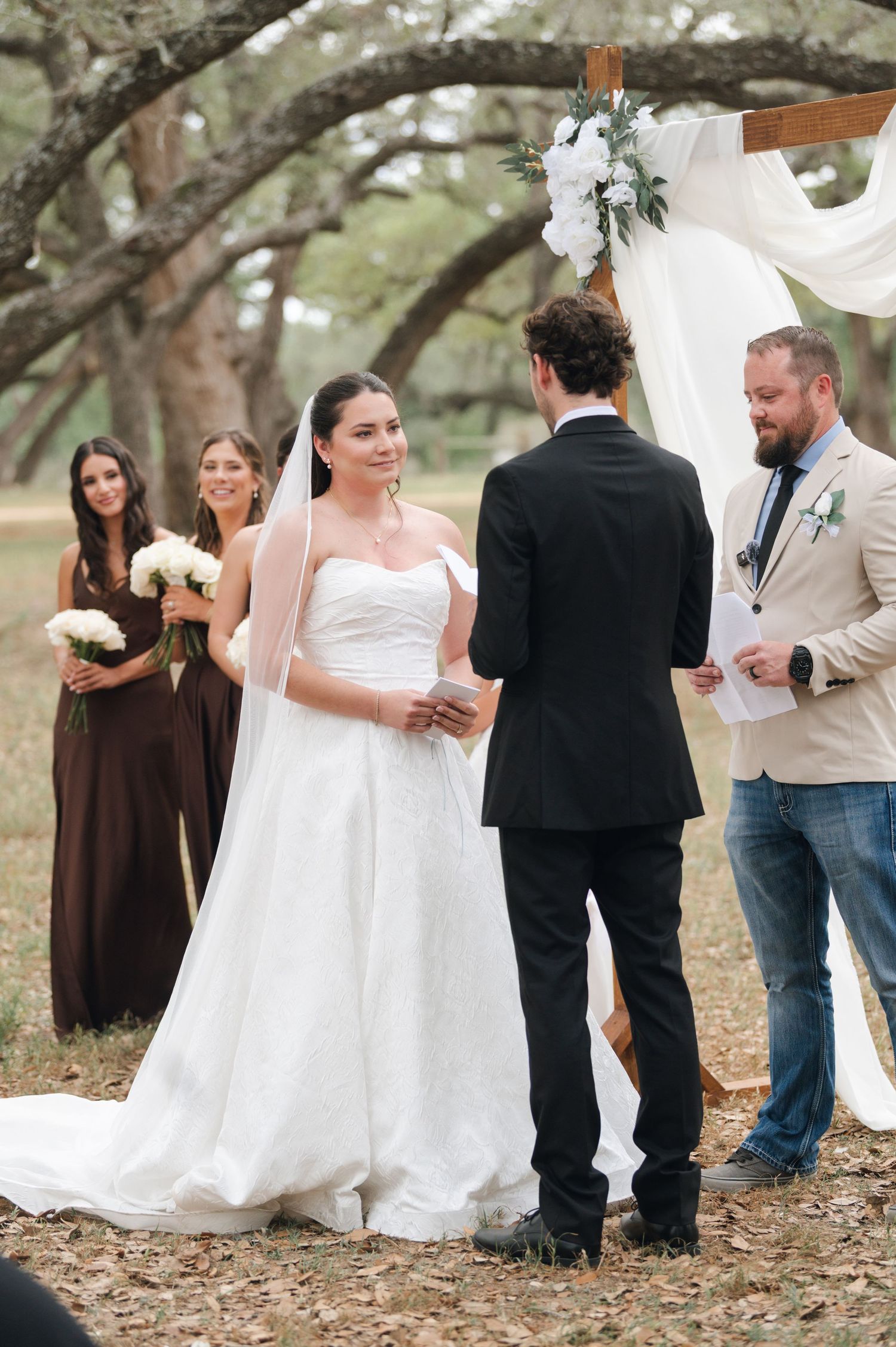 Bride smiling at groom during outdoor wedding ceremony under oak trees with bridesmaids in brown dresses.