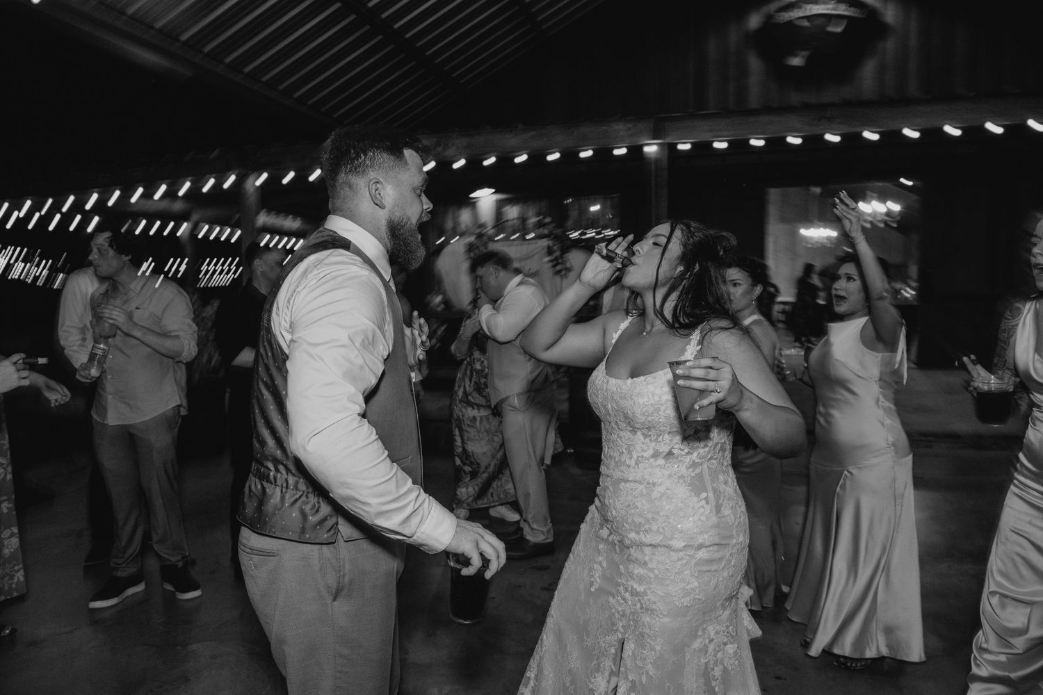 A bride and groom share a dance surrounded by guests at a lively wedding reception in black and white.