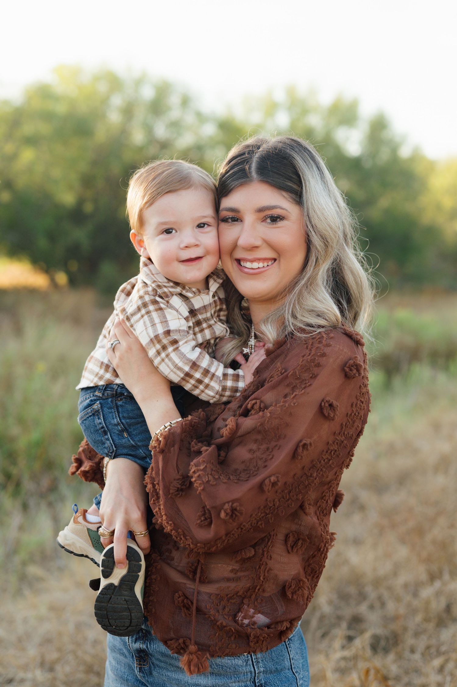 A parent and child share loving embraces in a golden field during sunset while wearing brown and plaid clothing.