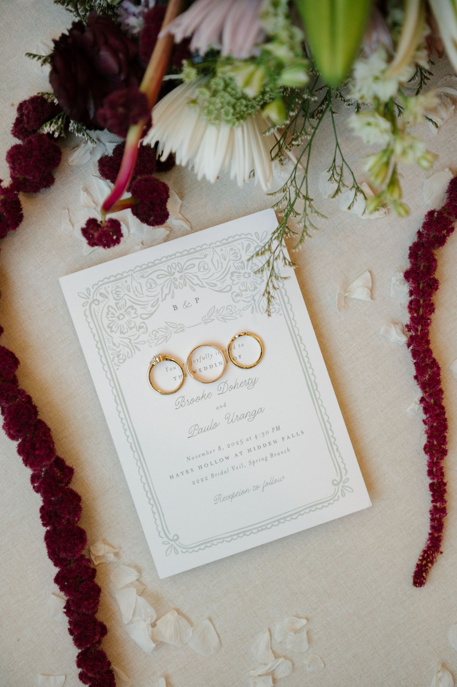Wedding rings and invitation displayed with burgundy florals and white stationery on light background.