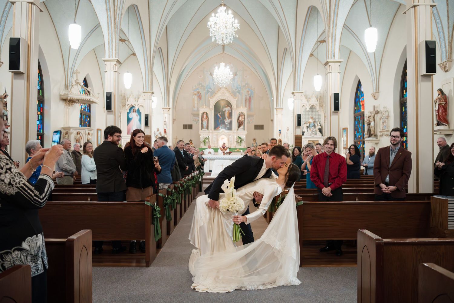Wedding ceremony taking place in a grand cathedral with ornate white arches and glowing chandeliers.