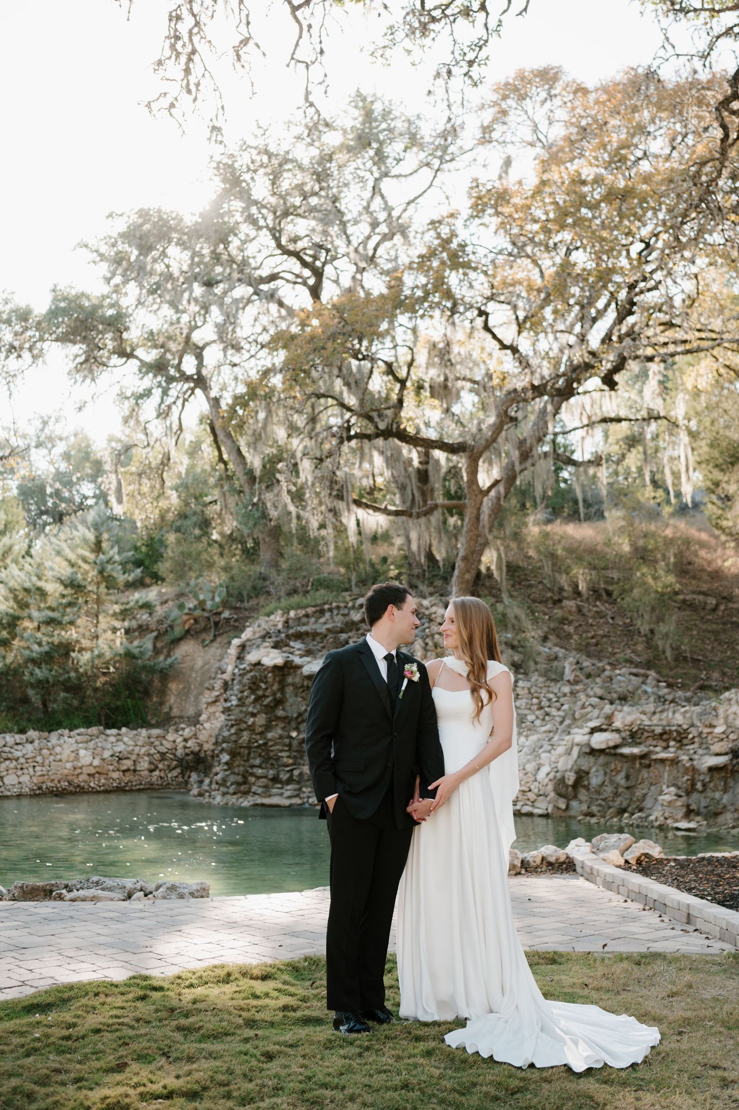 Bride and groom share intimate moment by scenic waterfront with Spanish moss draped trees.