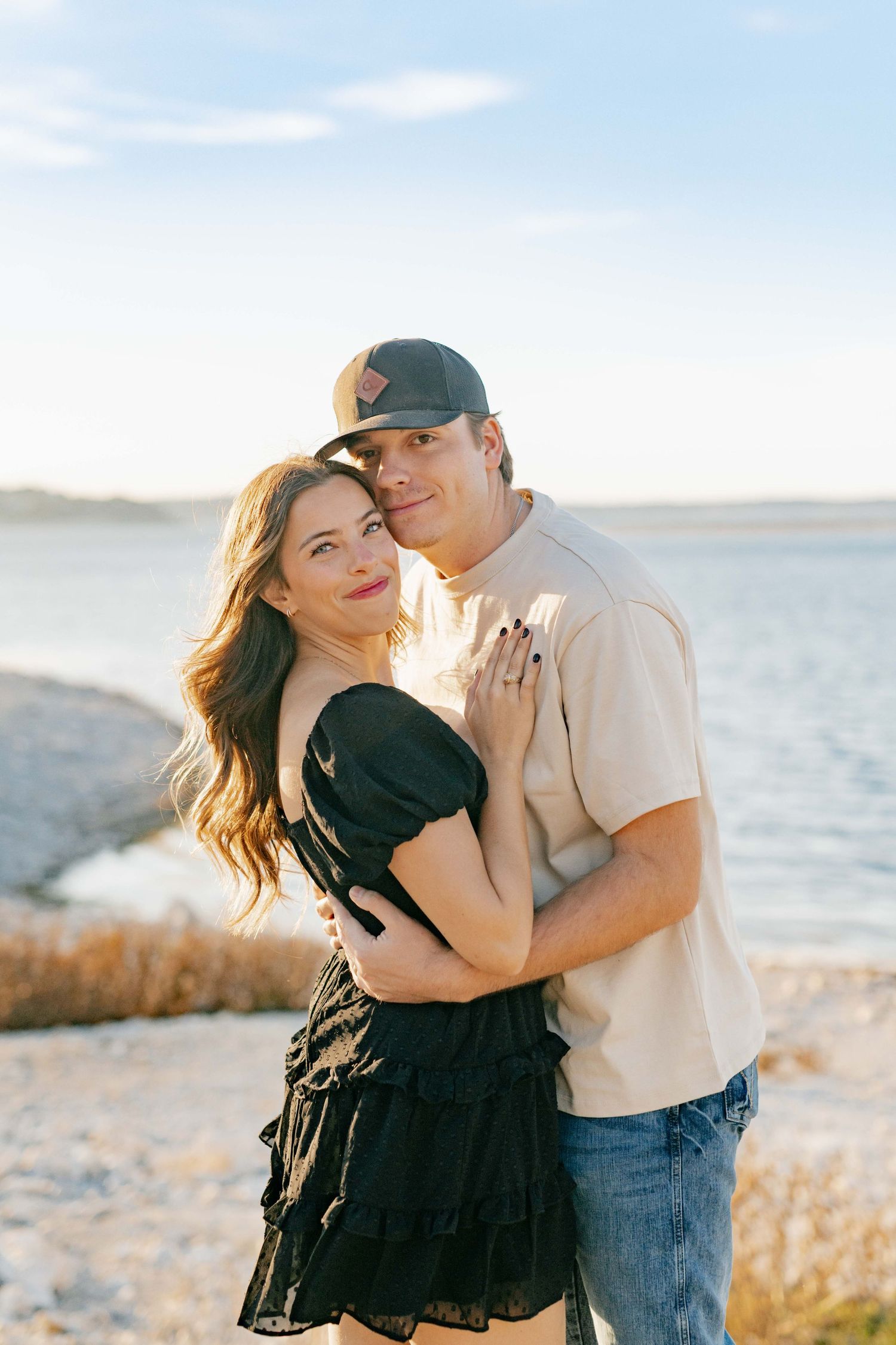 A romantic engagement photo of a couple in casual attire standing by a lake during golden hour.