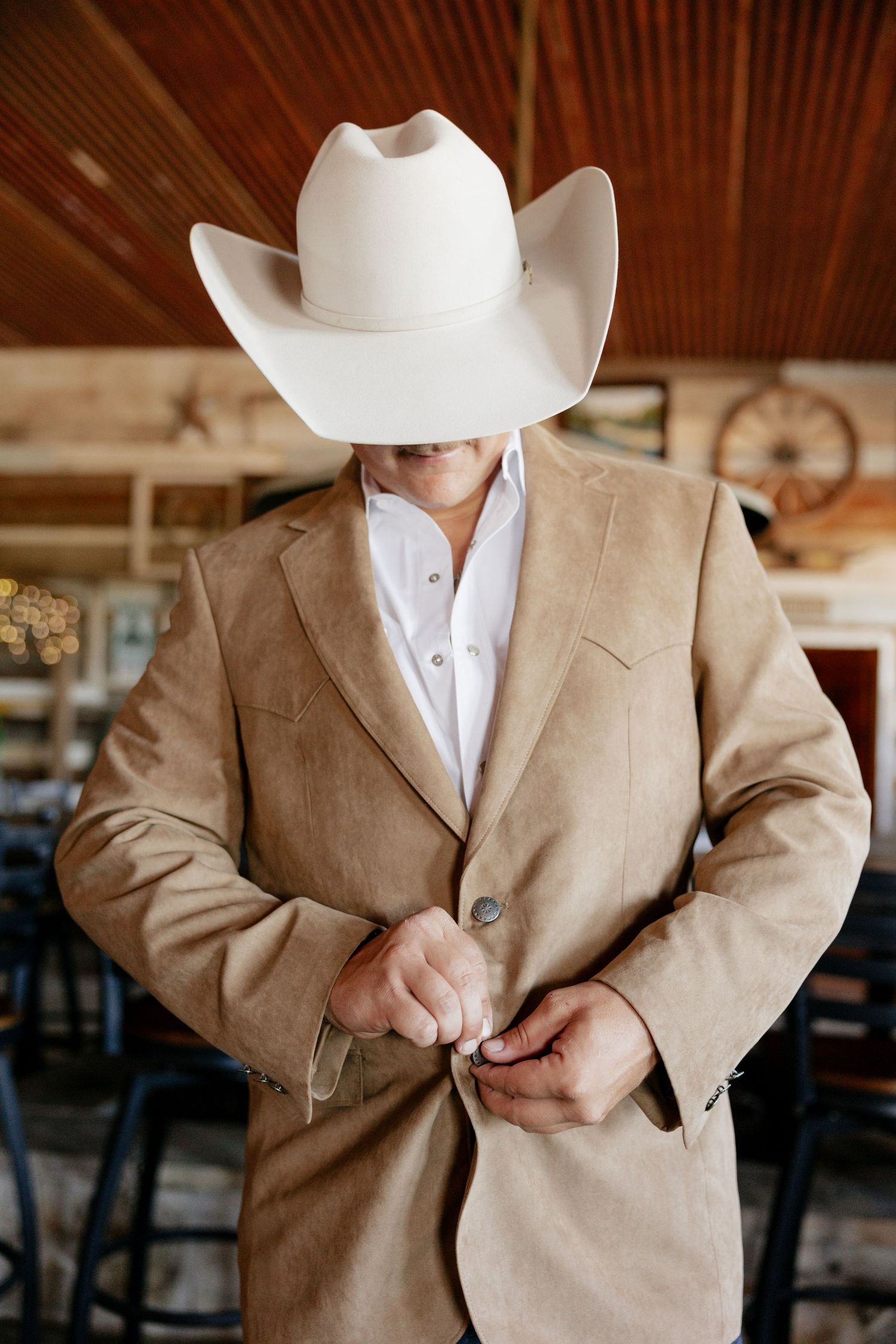 Western style groom in tan suit jacket and white cowboy hat buttoning up.