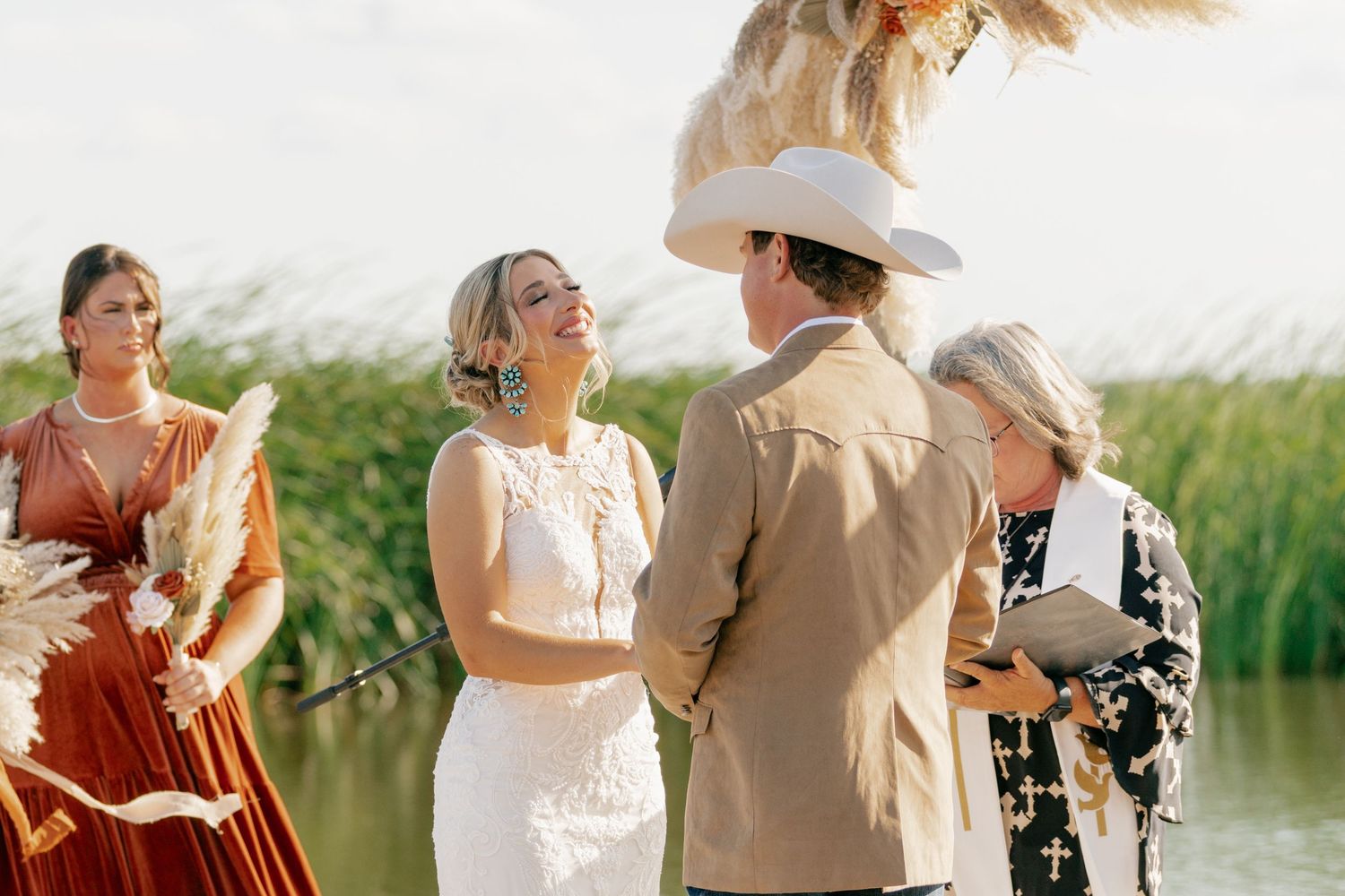 An outdoor wedding ceremony takes place by a scenic lakeside with tall grass and natural surroundings in warm sunlight.