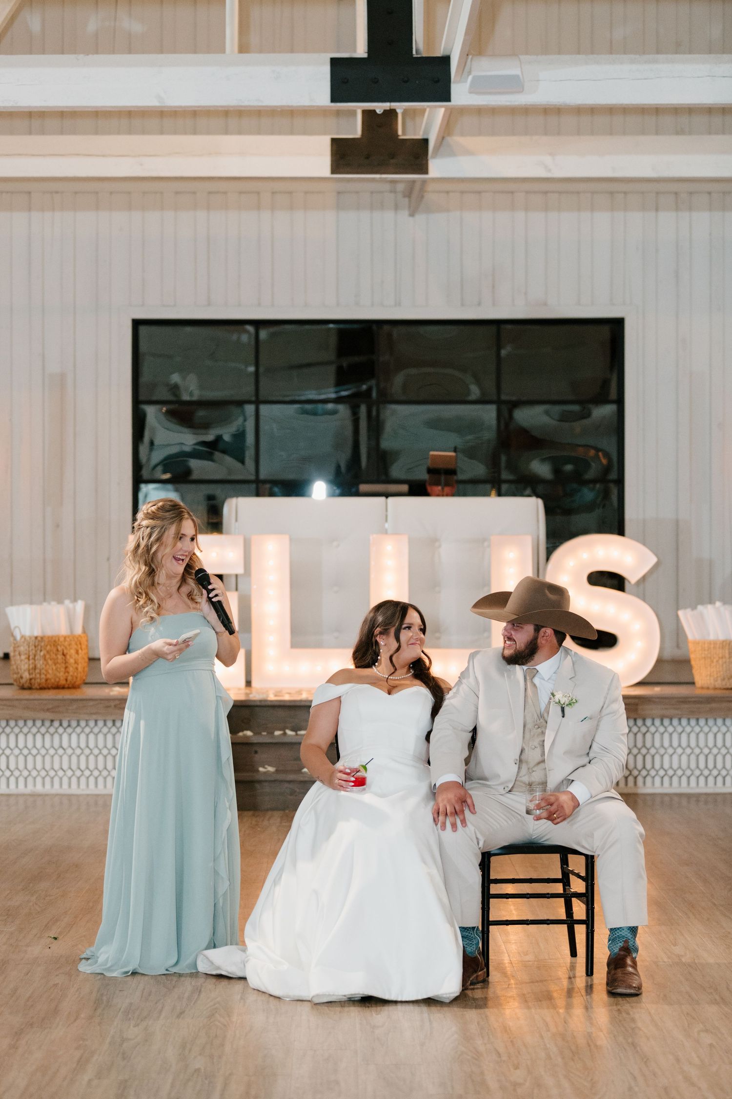 A couple at their wedding reception sits in front of illuminated letters with a bridesmaid in a light blue dress nearby.