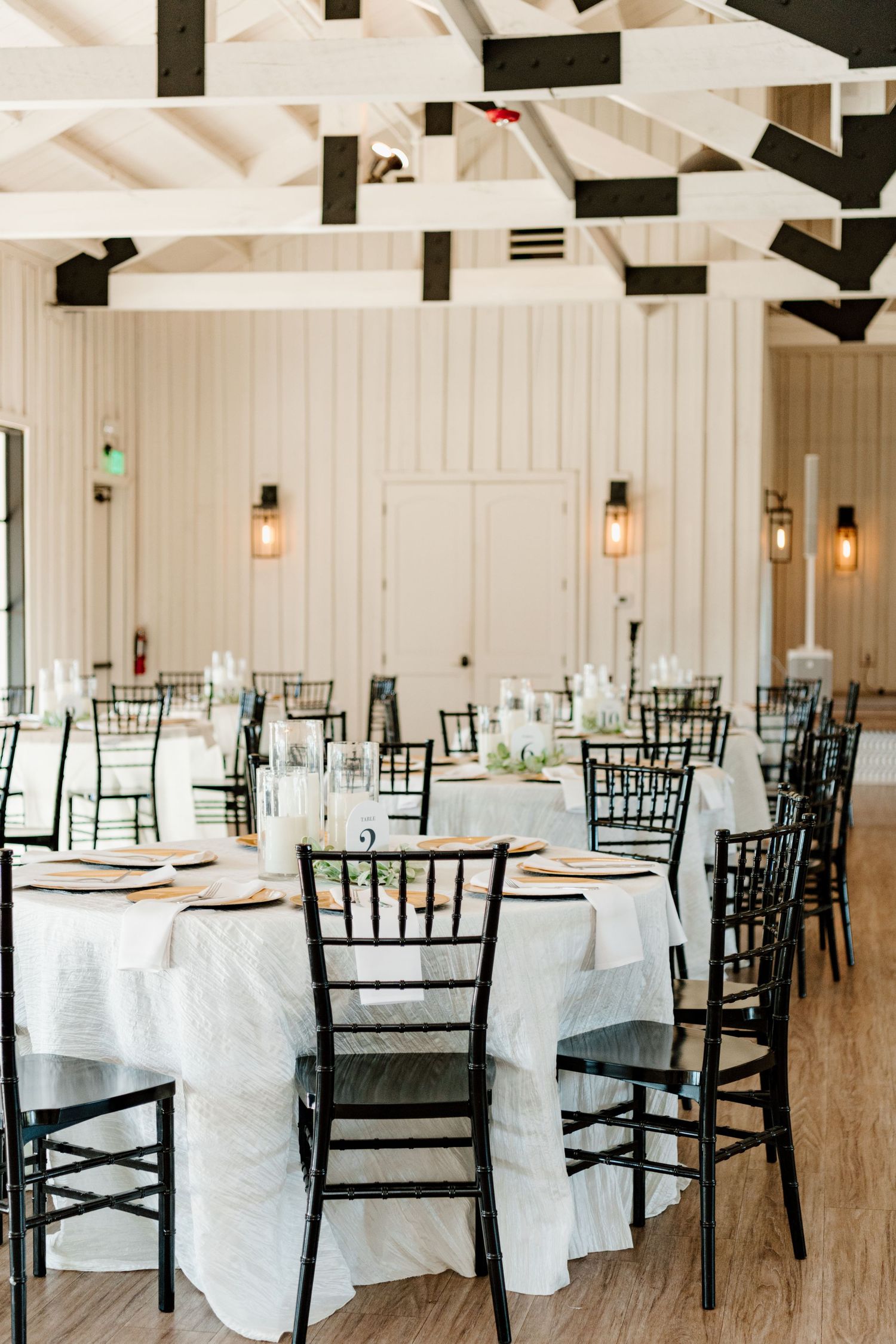 Elegant wedding reception venue with black Chiavari chairs and white tablecloths under exposed beam ceilings with pendant lights.