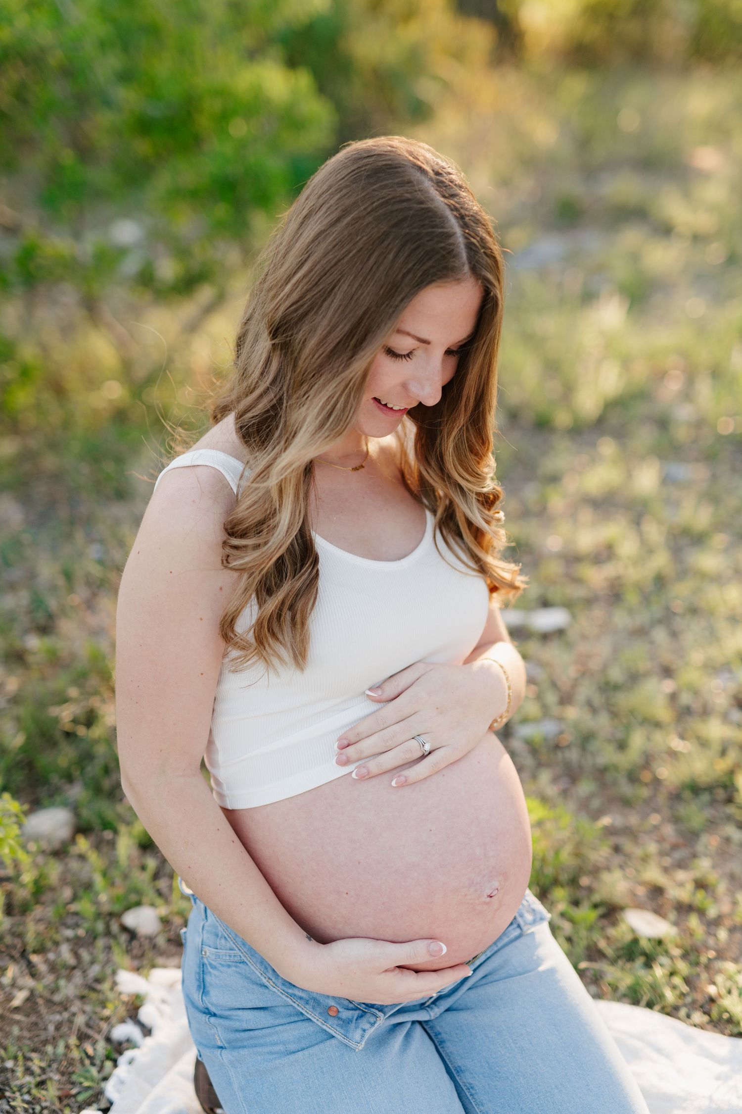 A pregnant woman in a white top and jeans sits on a blanket outdoors, gently cradling her baby bump in natural lighting.