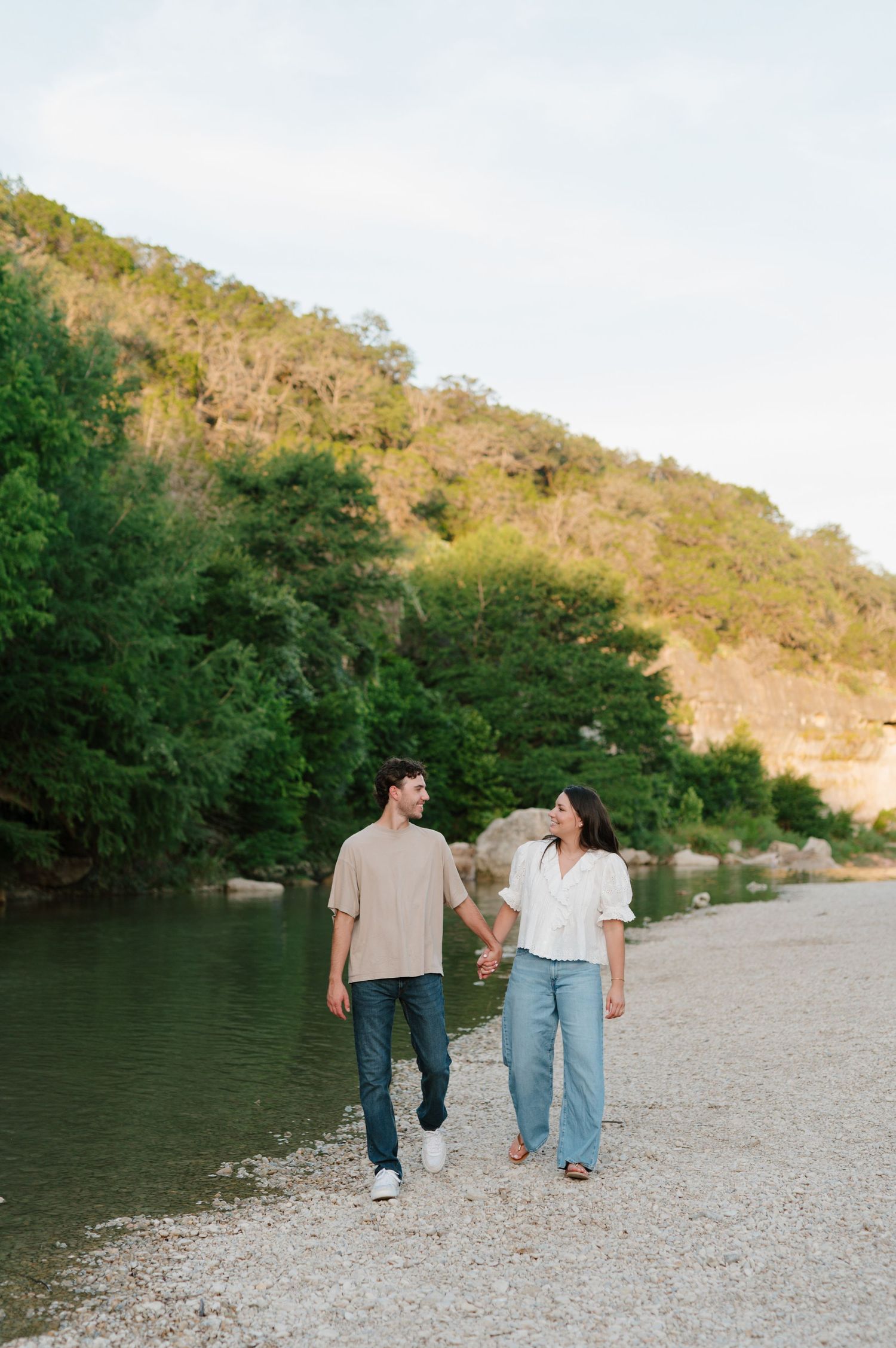 A couple walks hand in hand along a rocky shoreline with hills and trees in the background.