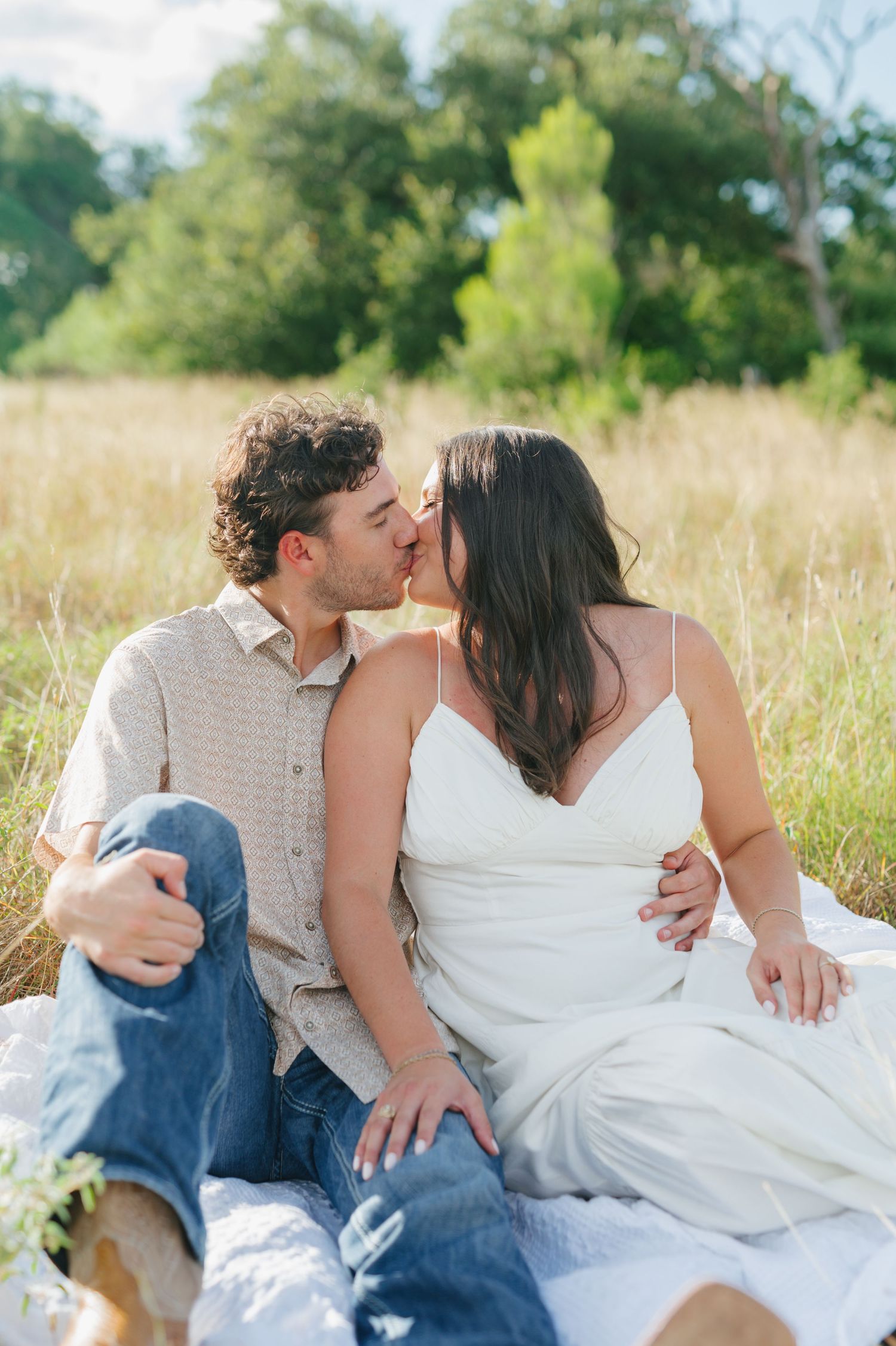 A couple sharing an intimate moment while sitting in a sunlit meadow with tall grass.