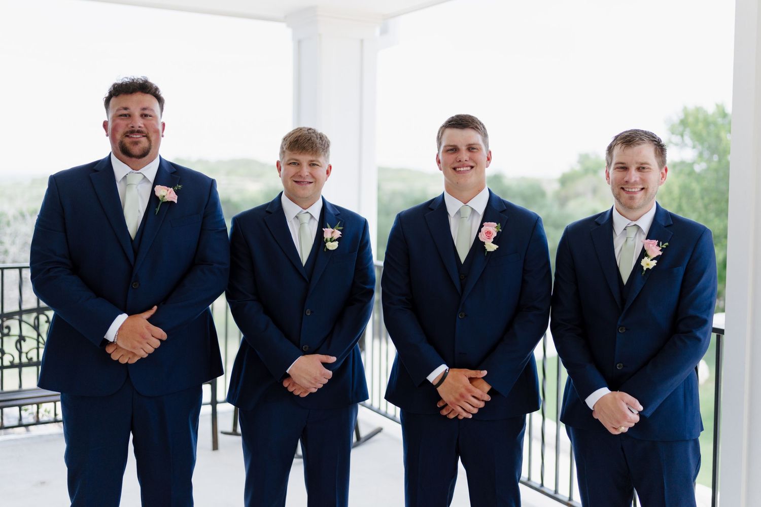 Groomsmen line up wearing matching navy suits with boutonnieres against a white backdrop.