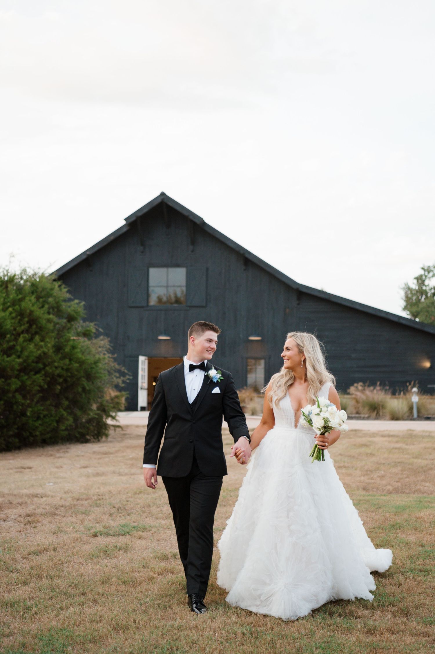 A bride and groom walking together on a grassy field in front of a rustic barn.