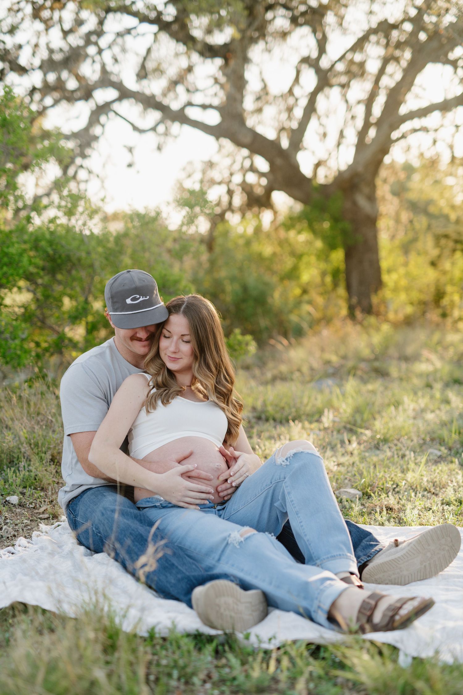 A couple sits on a white blanket sharing an intimate moment during their maternity photoshoot.