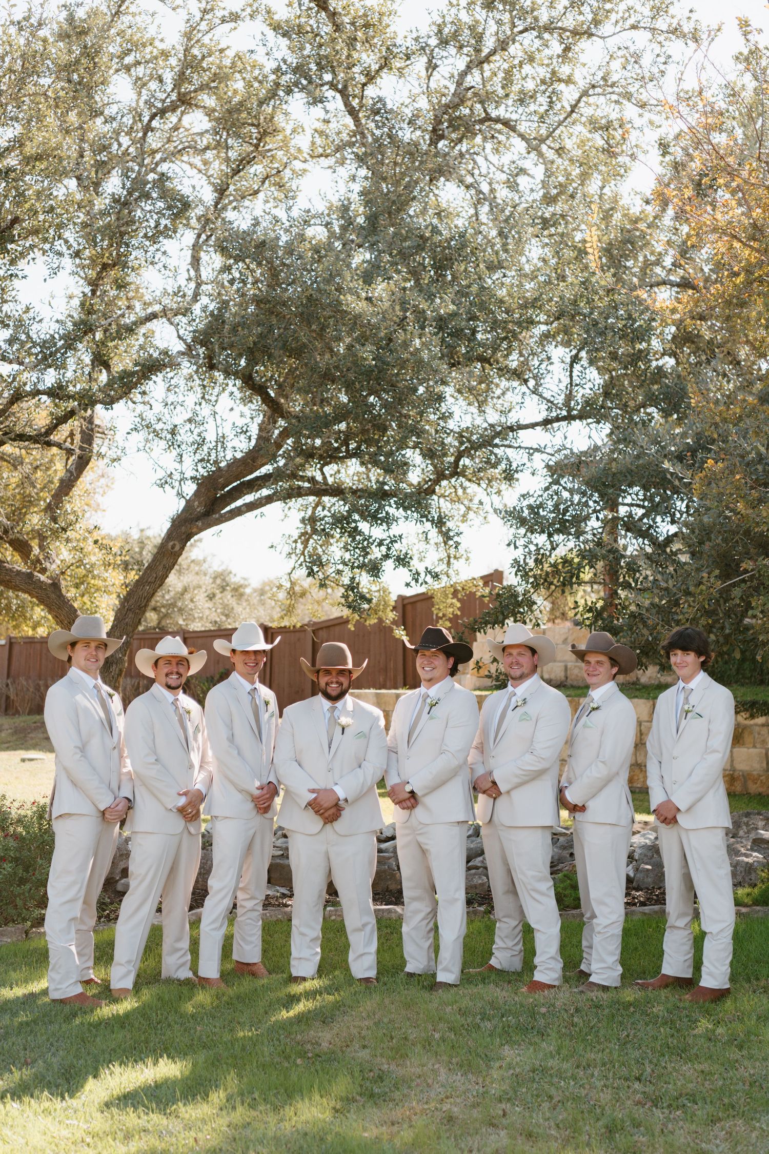 A group in matching white suits and cowboy hats poses together outdoors under a large tree.