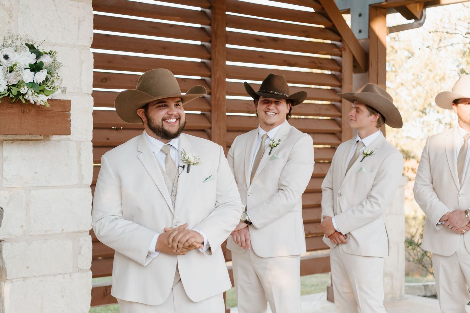 Three groomsmen in light beige suits and cowboy hats stand together at a rustic outdoor wedding ceremony venue.