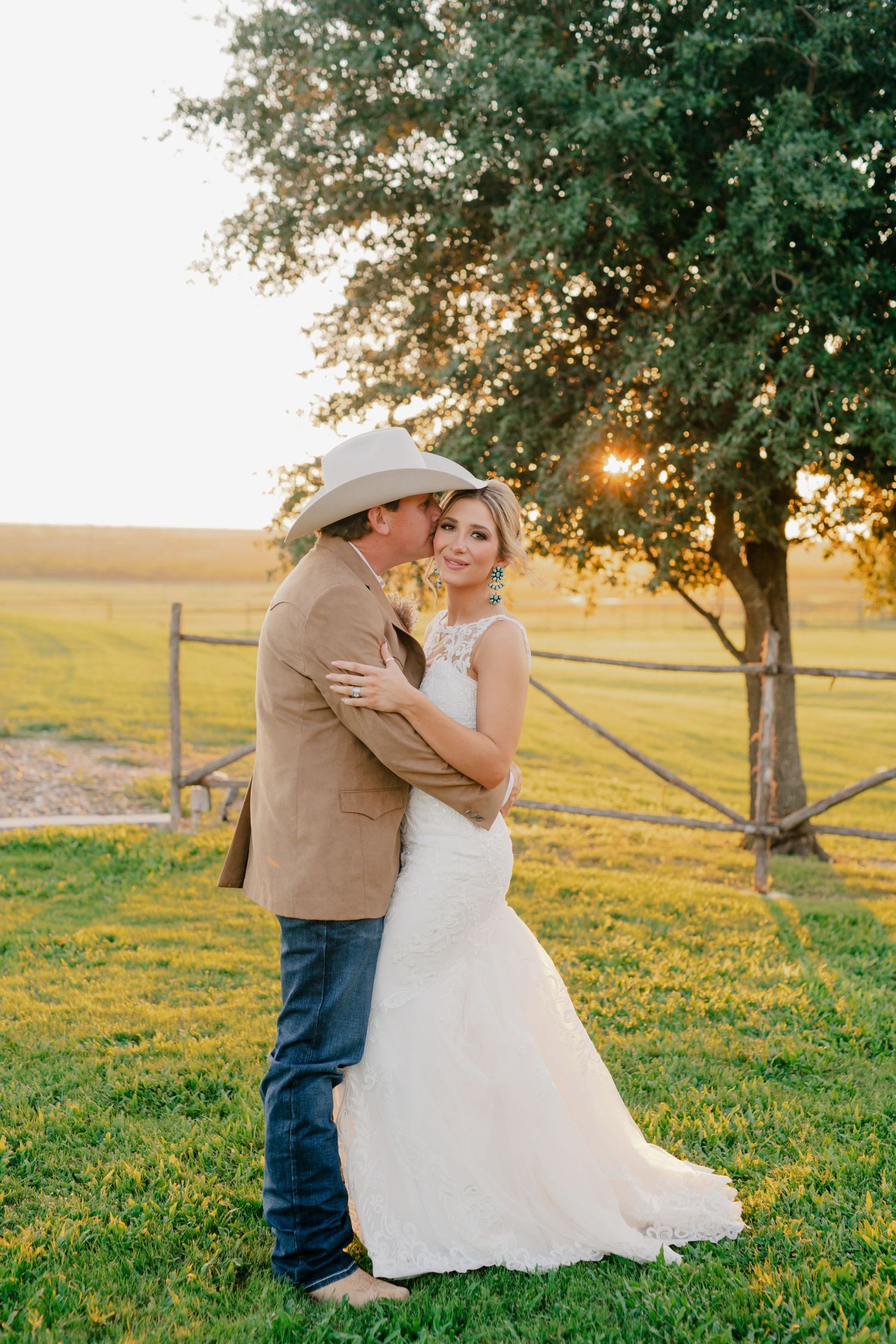 Newlyweds embracing under tree at sunset in rustic countryside setting.