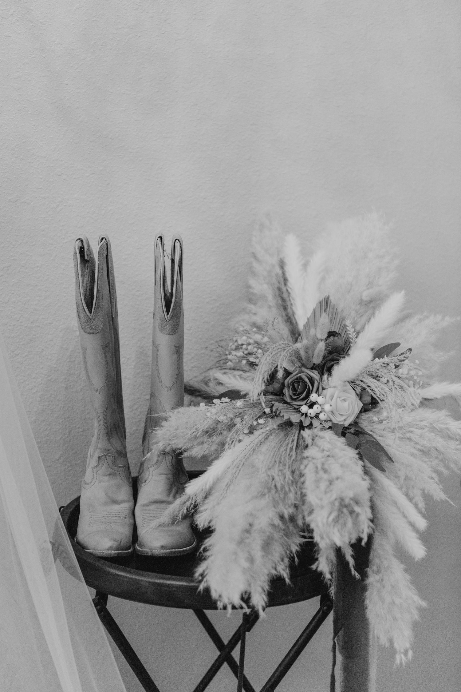 A pair of beige cowboy boots and a white floral bouquet with pampas grass displayed on a black folding chair.