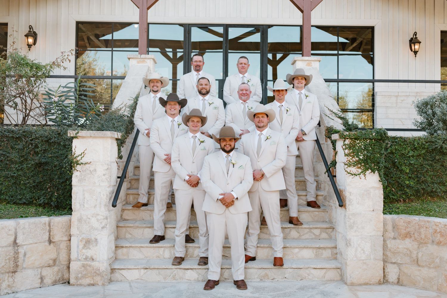A wedding party in all-white attire poses on stone steps in front of a rustic venue.