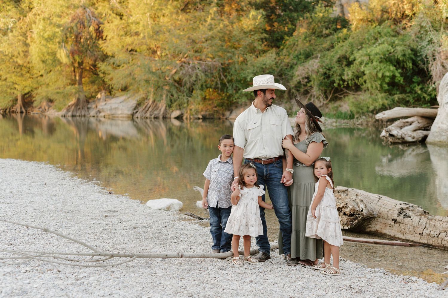 A family group stands together on a rocky riverbank surrounded by autumn foliage and calm water.