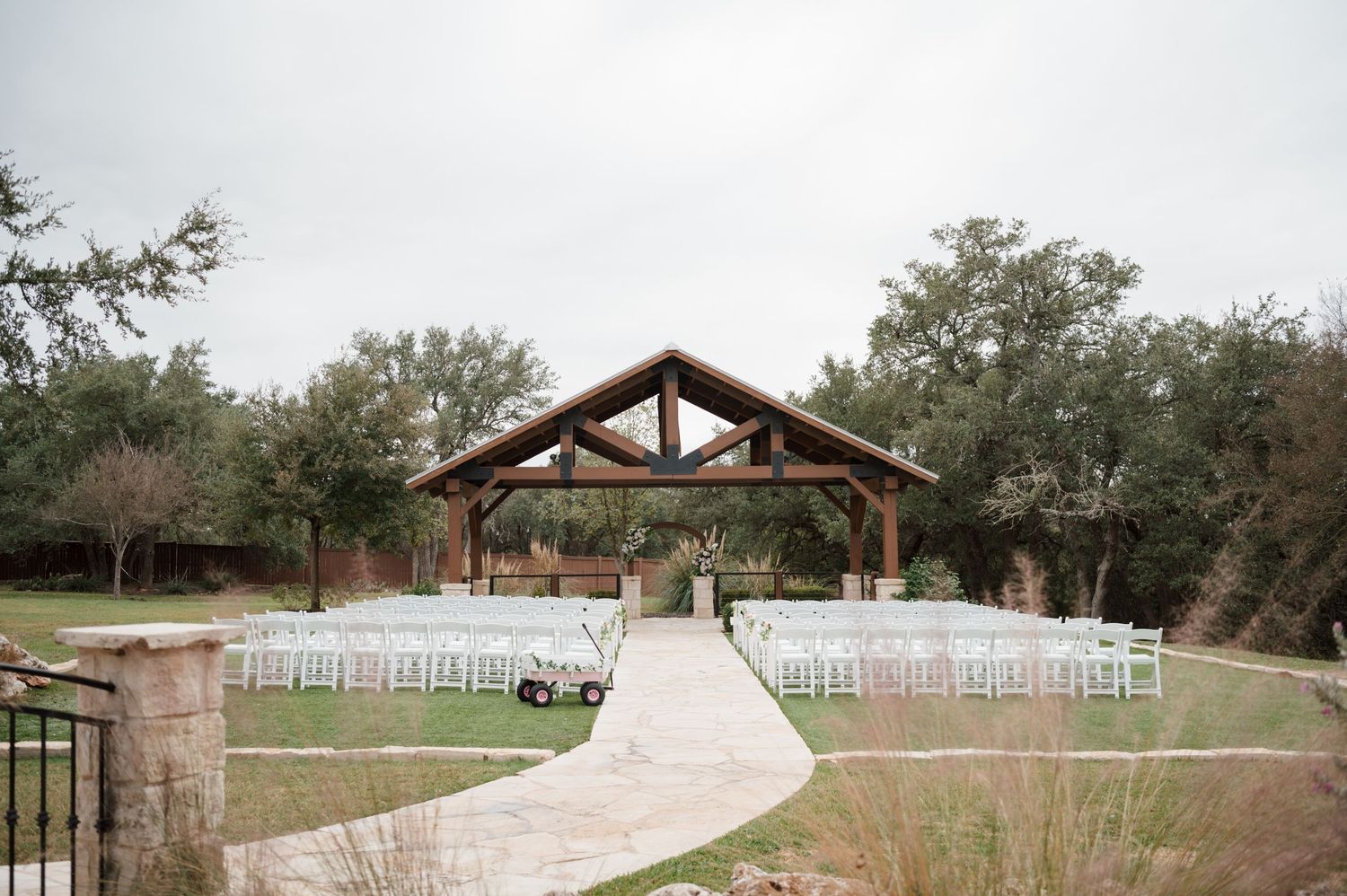 A scenic outdoor wedding venue features a stone path leading to a wooden pergola surrounded by lush greenery and white chairs.