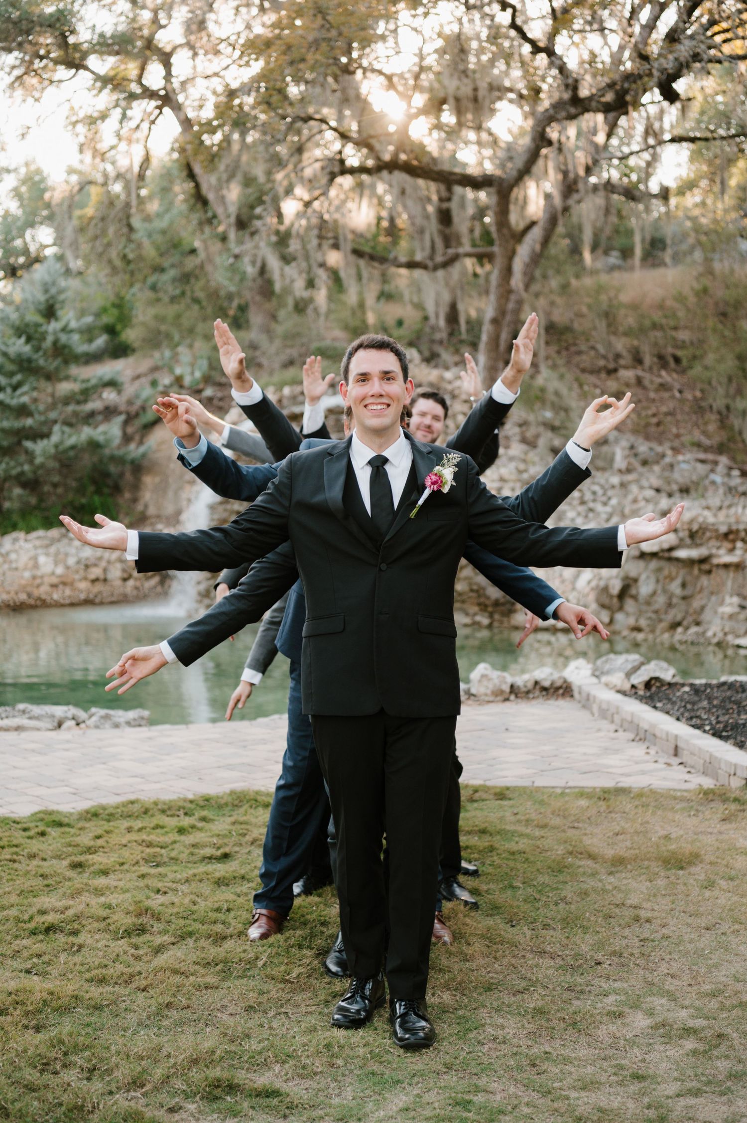 Groomsmen in black suits strike playful poses with outstretched arms by a waterfront setting.