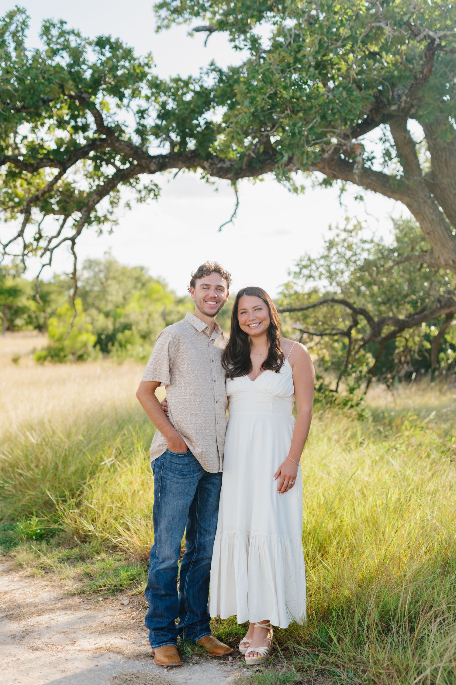 Couple poses together in a sunny field with tall grass and trees in the background.