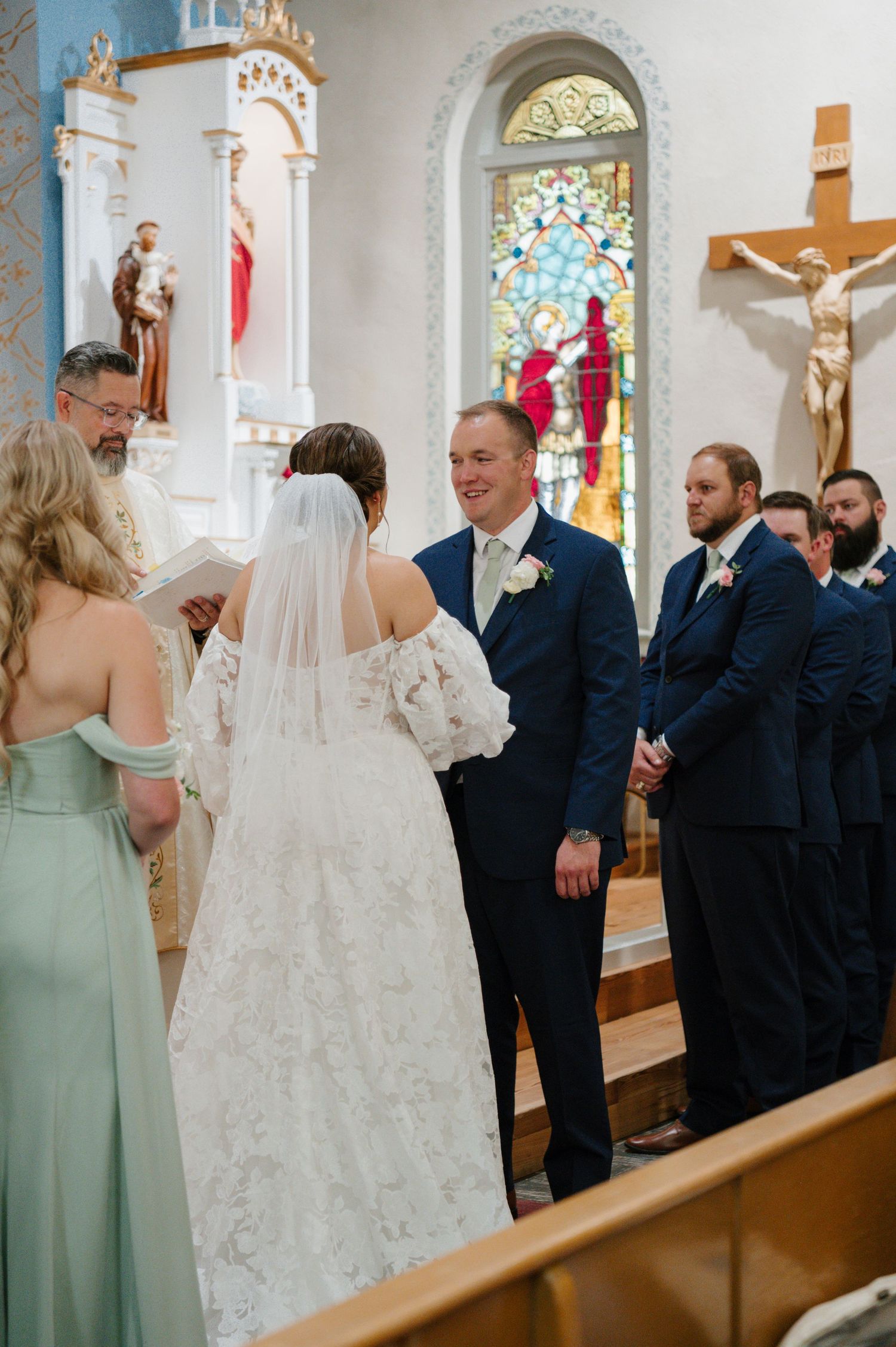 Church wedding ceremony with bridesmaids in sage green dresses and groomsmen in navy suits.