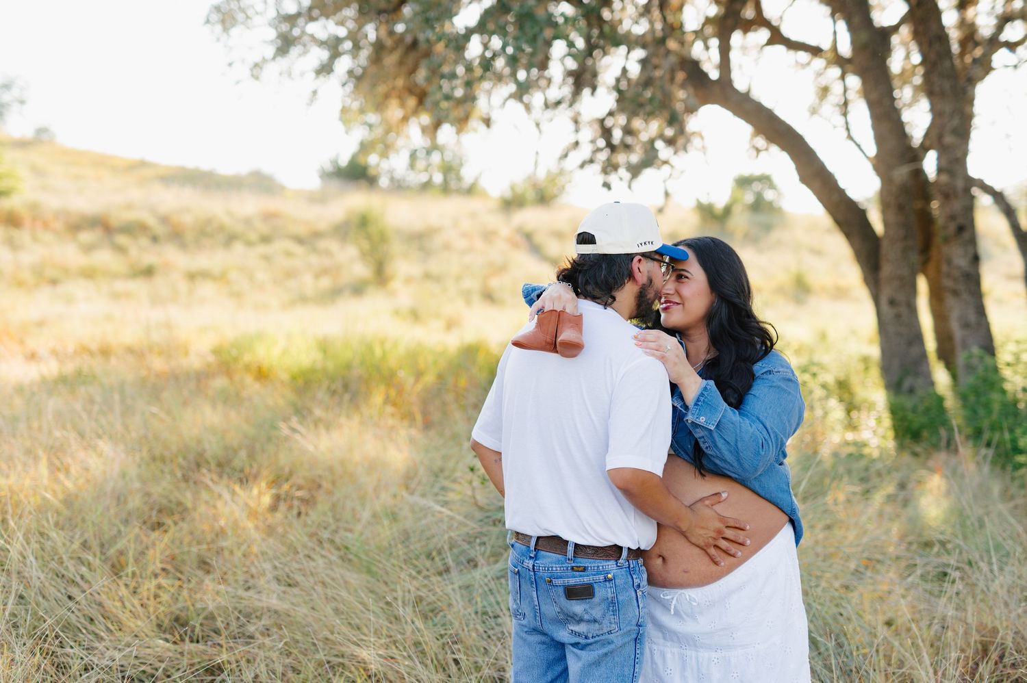 A couple in blue denim and white outfits embraces tenderly in a sun-dappled natural setting.